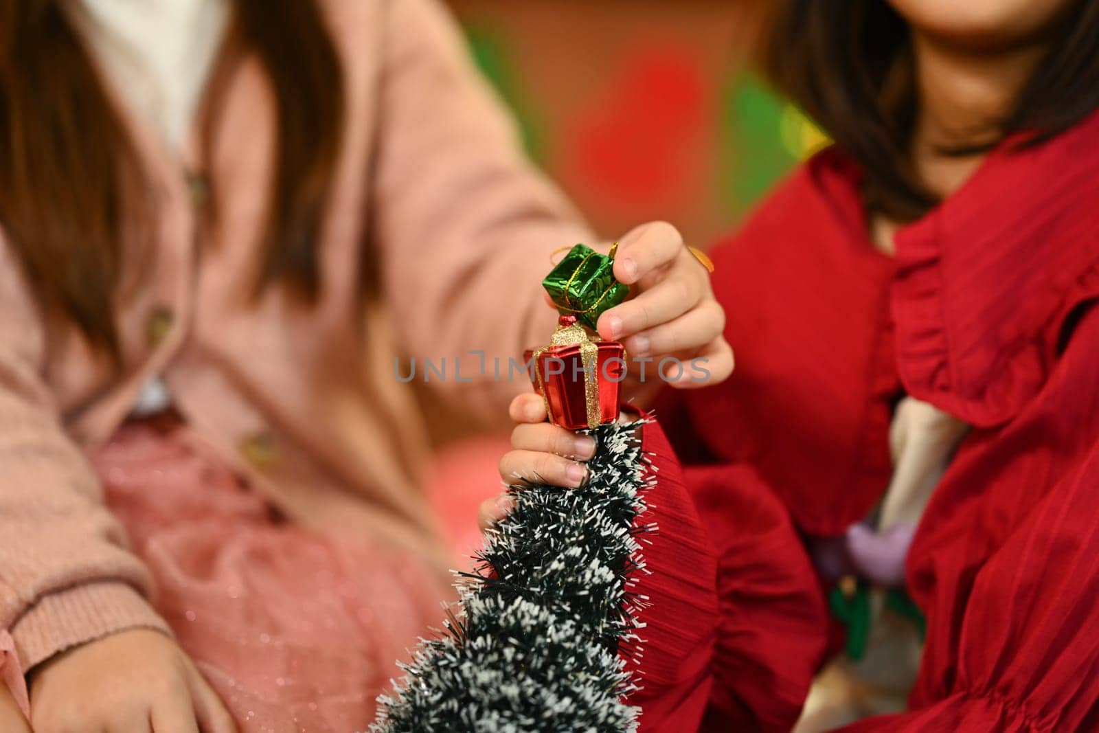Two little children wearing warm winter clothes decorating small Christmas tree in living room.