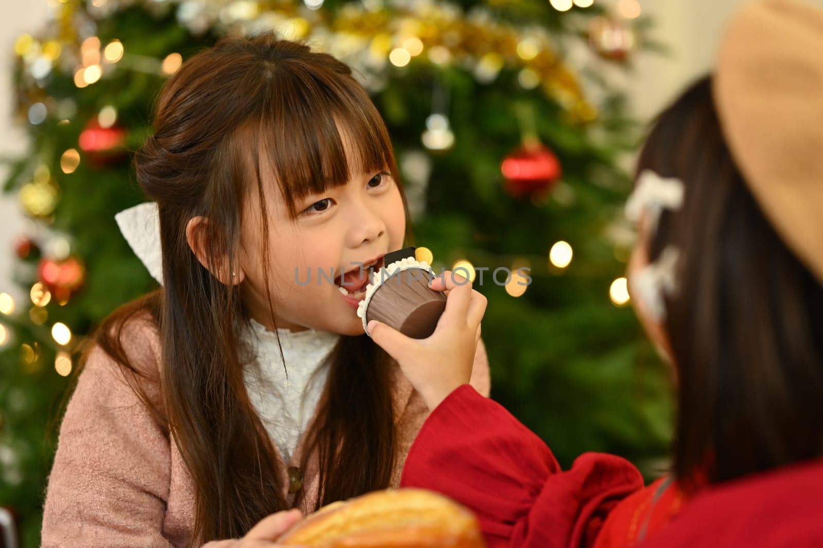 Cute little girl eating cupcake with a beautifully decorated Christmas tree on background by prathanchorruangsak