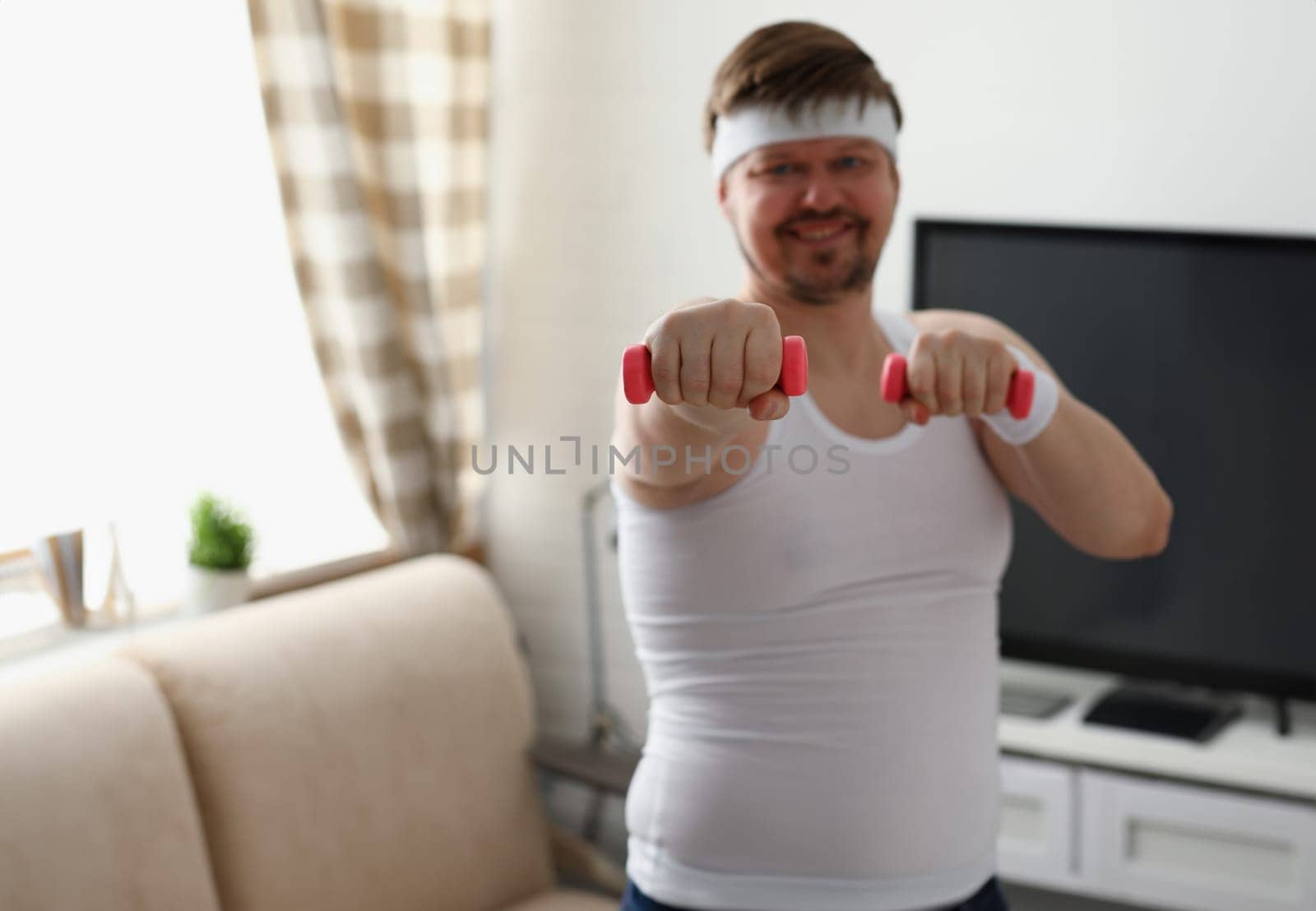 Young attractive man engaged in fitness at home holding dumbbell in hand trying to fight excess weight and loss