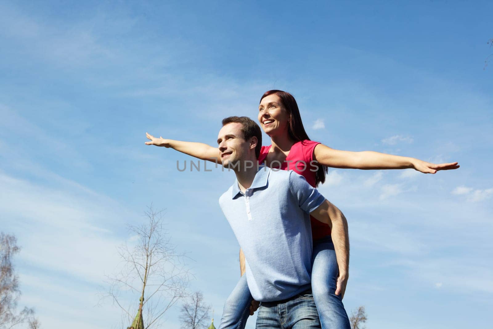 Happy man giving piggyback to his wife outdoors against blue sky