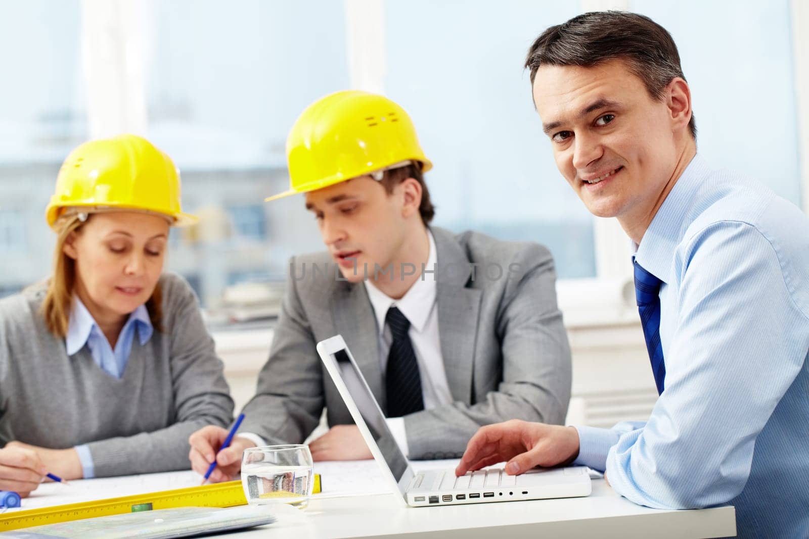 A manager working with laptop, looking at camera and smiling against his colleagues architects