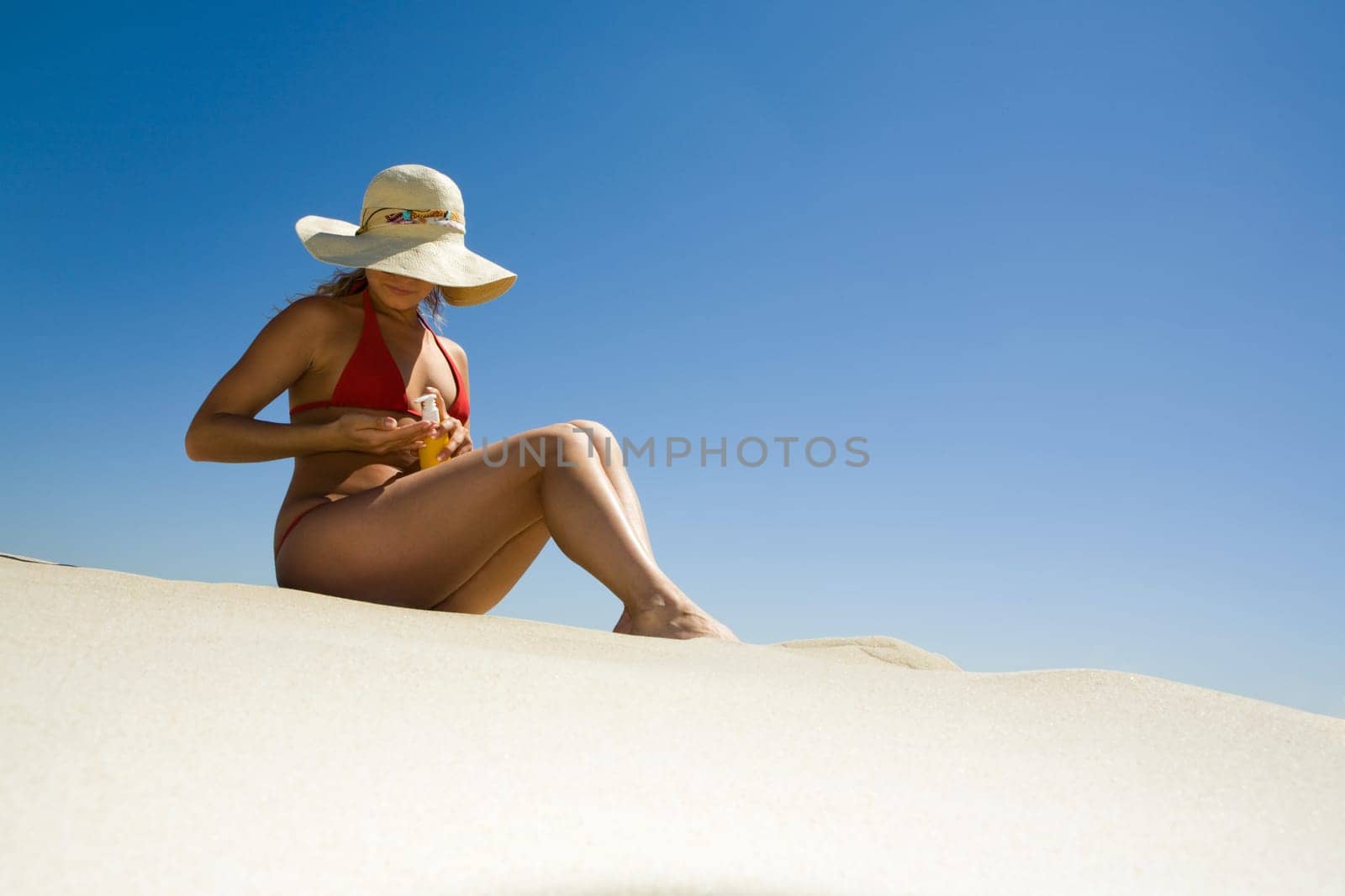 Image of pretty woman in hat sitting on sand and applying body lotion before sunbathing
