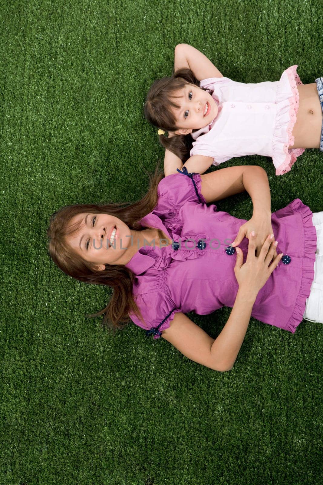 Happy child and her mother lying on green grassland and looking at camera with smiles