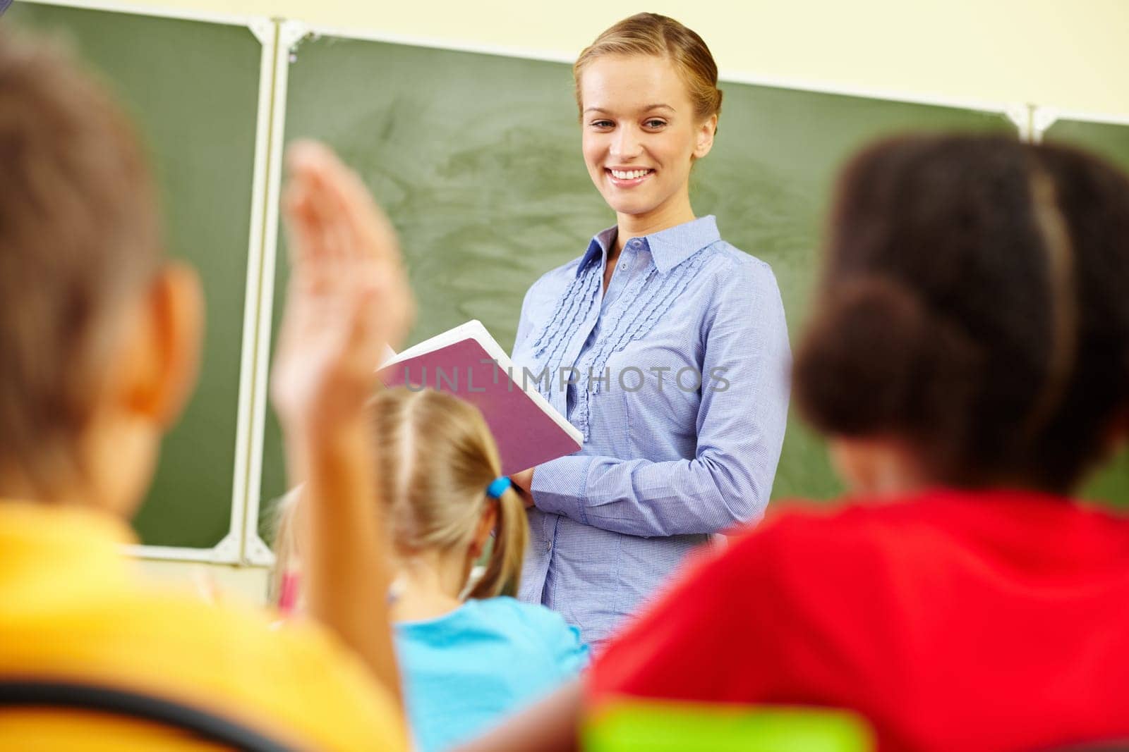 Portrait of smart teacher with exercise book looking at schoolkids in classroom