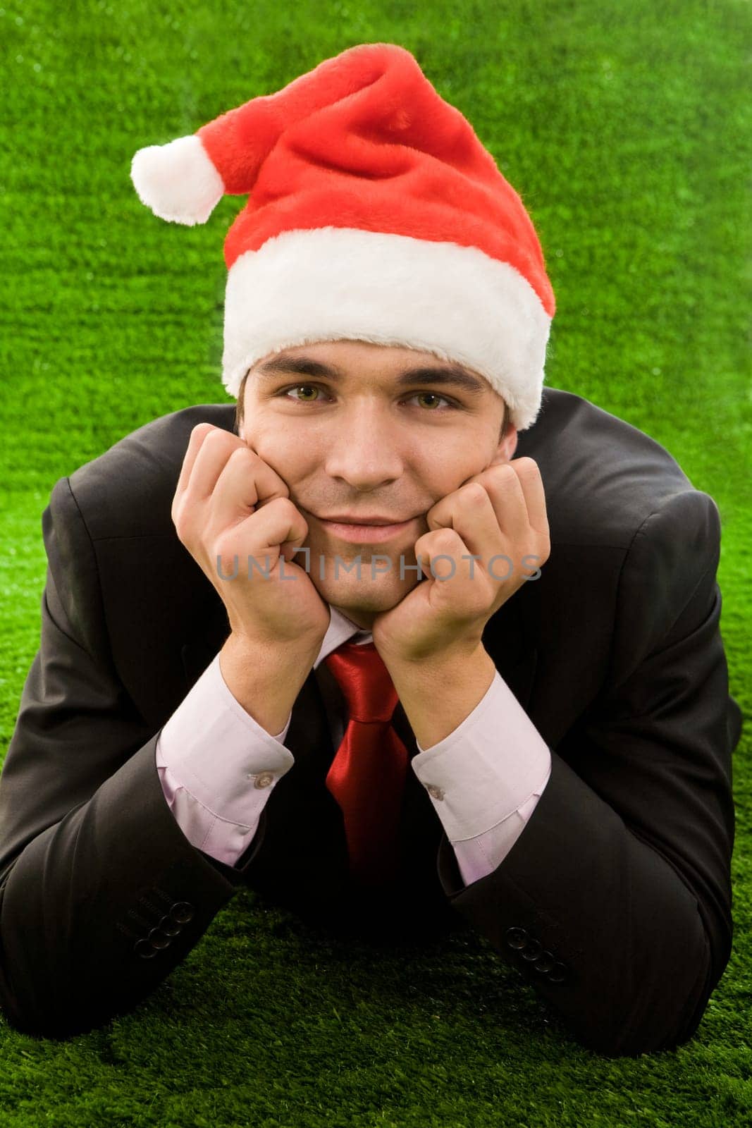 Businessman in santa cap touching his face by hands while relaxing on green grass