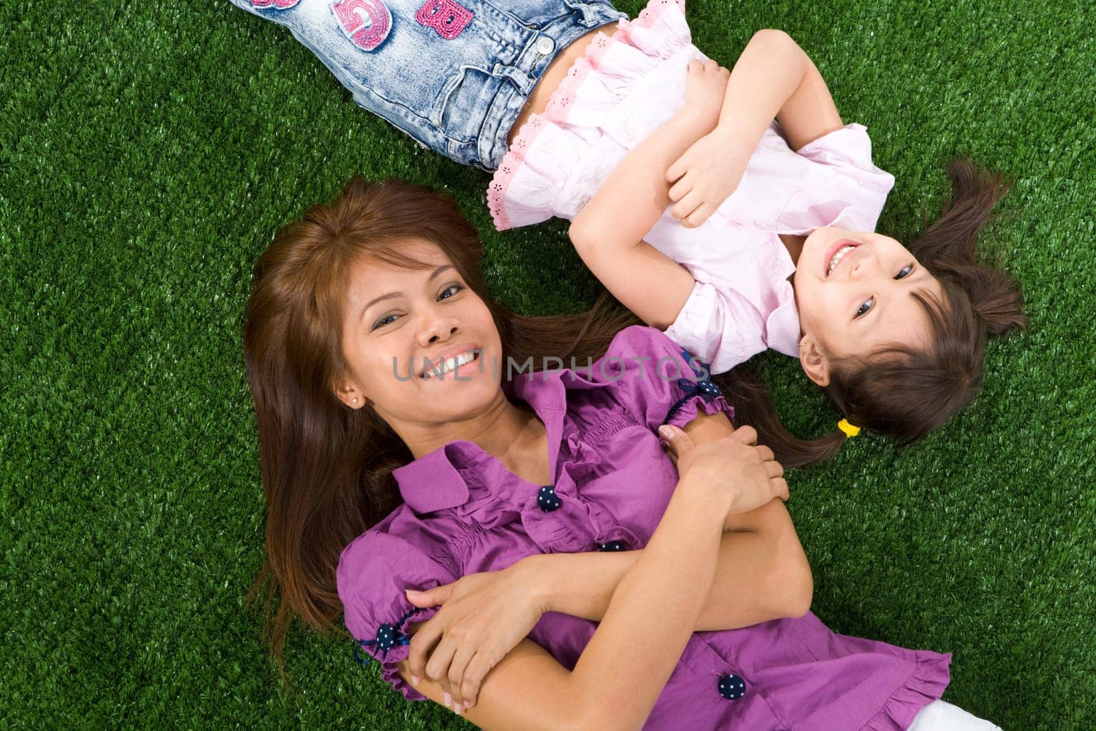 Happy little girl and her mother lying head to head on green grass and looking at camera