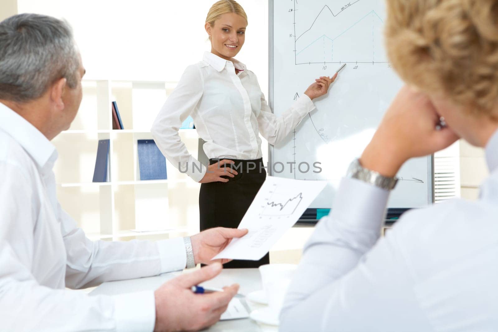 Photo of successful employee standing by whiteboard while her colleagues listening to her
