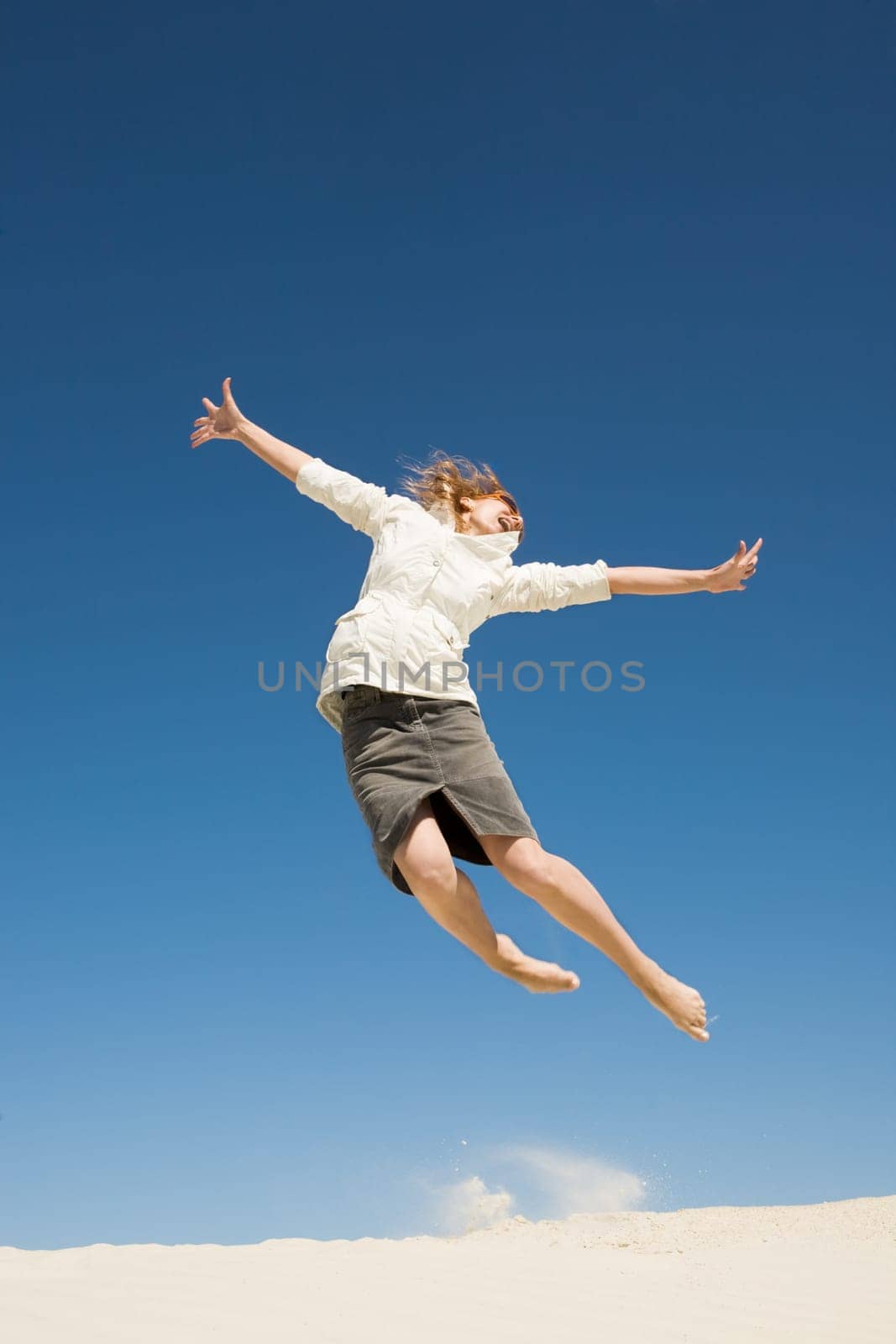 Photo of joyful female in casual clothes jumping over sand with her arms outstretched