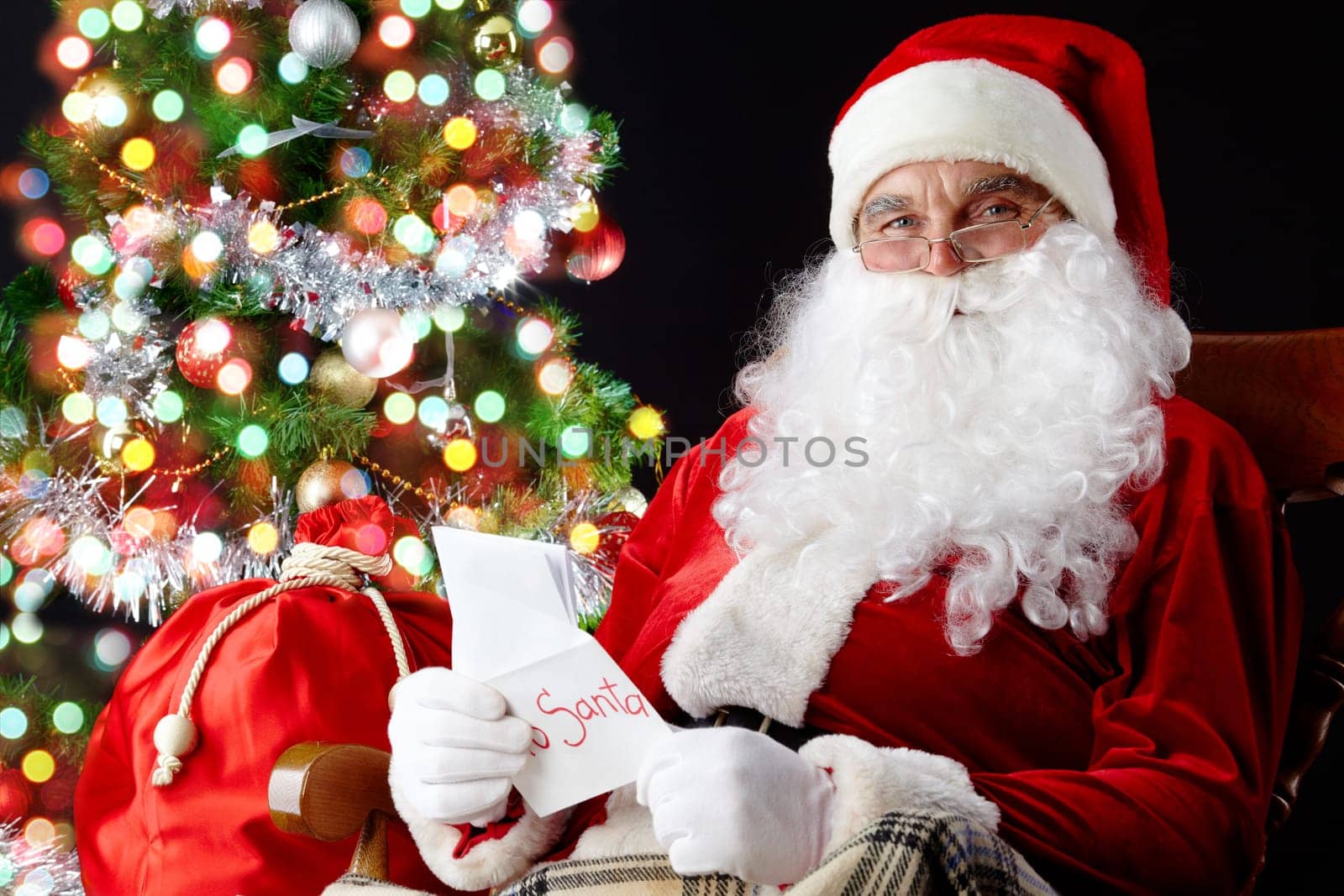 Santa sitting at the Christmas tree, holding Christmas letters and looking at camera