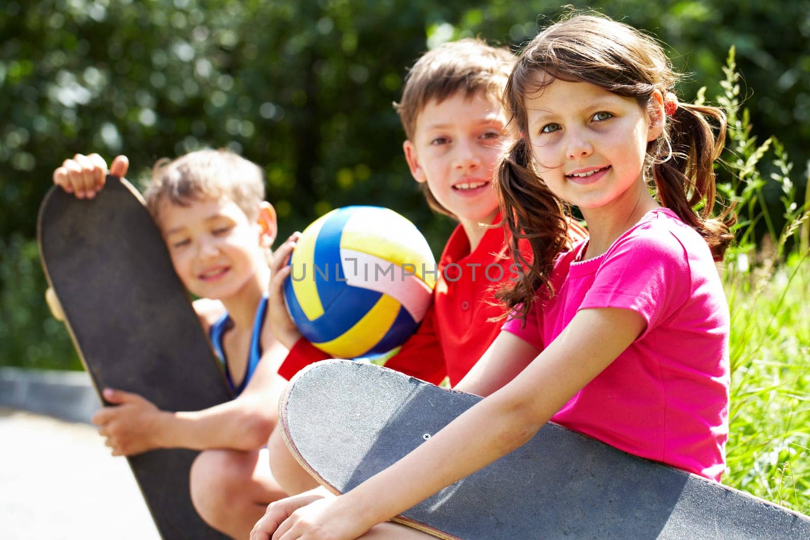 Three little children with skateboards sitting on asphalt, looking at camera and smiling