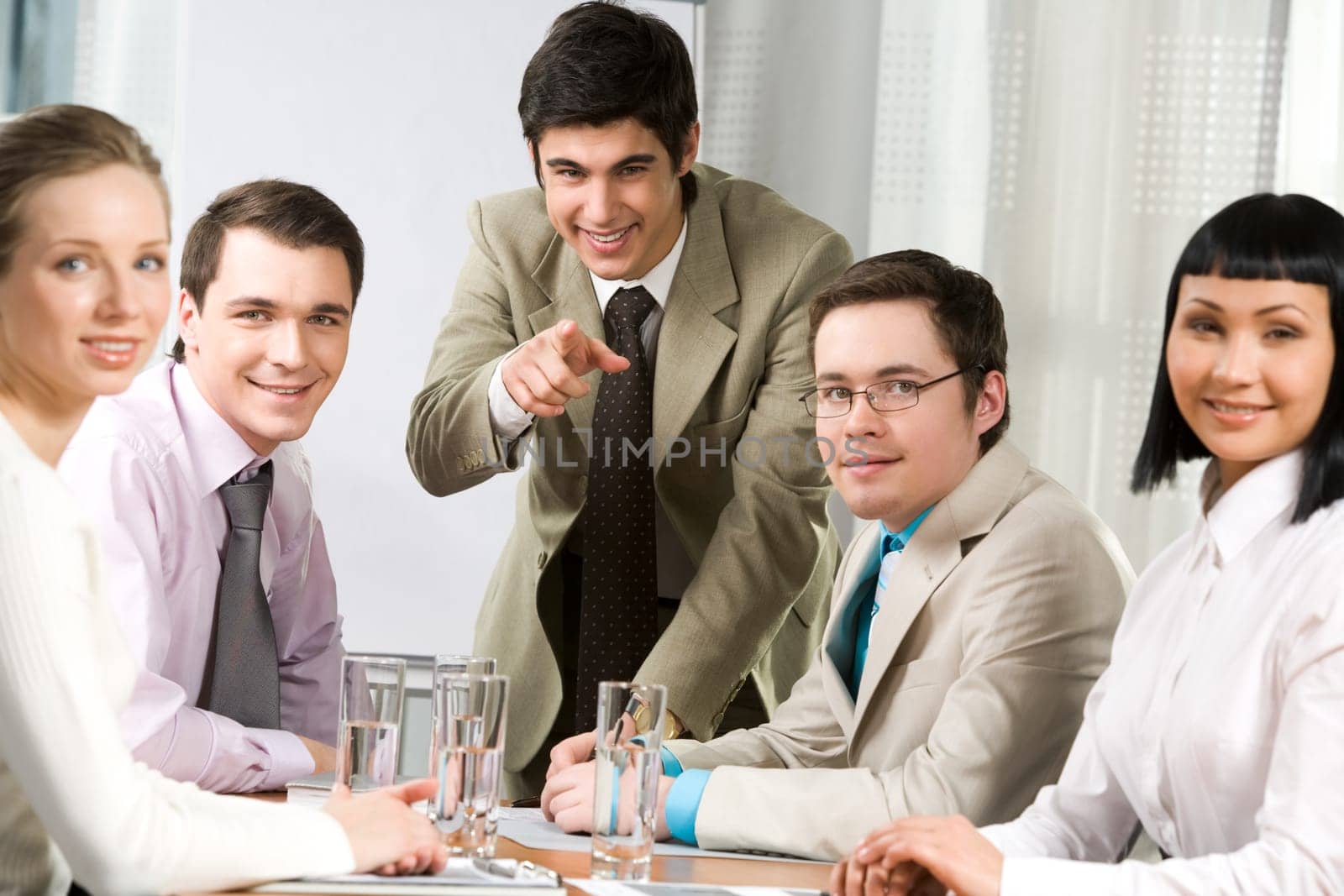 Image of friendly workteam sitting around table with their leader pointing at camera