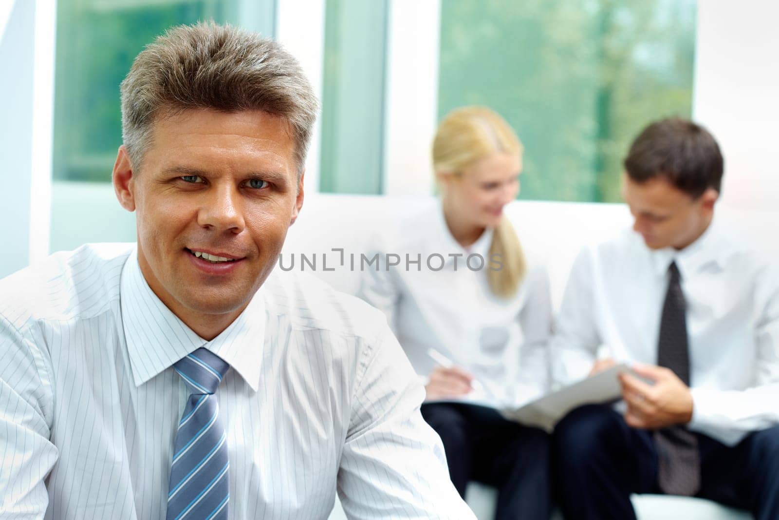Confident businessman looking at camera at background of his communicating colleagues