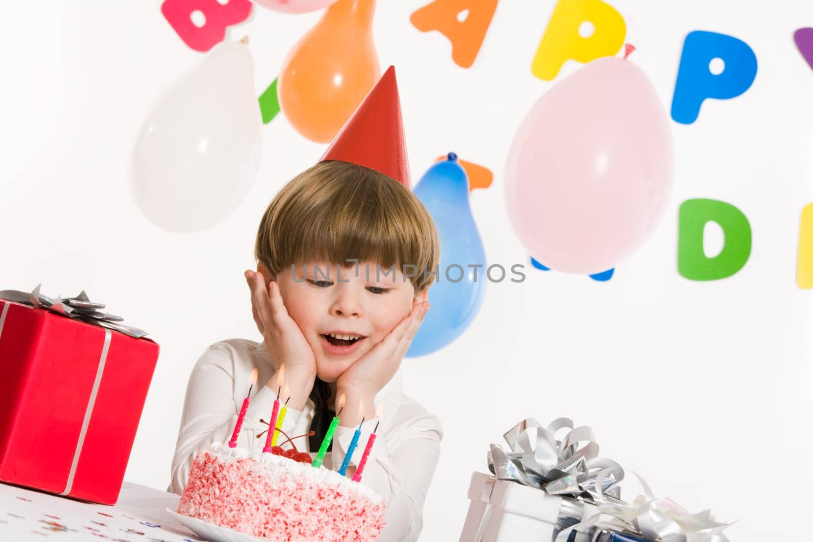 Portrait of surprised lad touching his cheeks and looking at burning candles on birthday cake