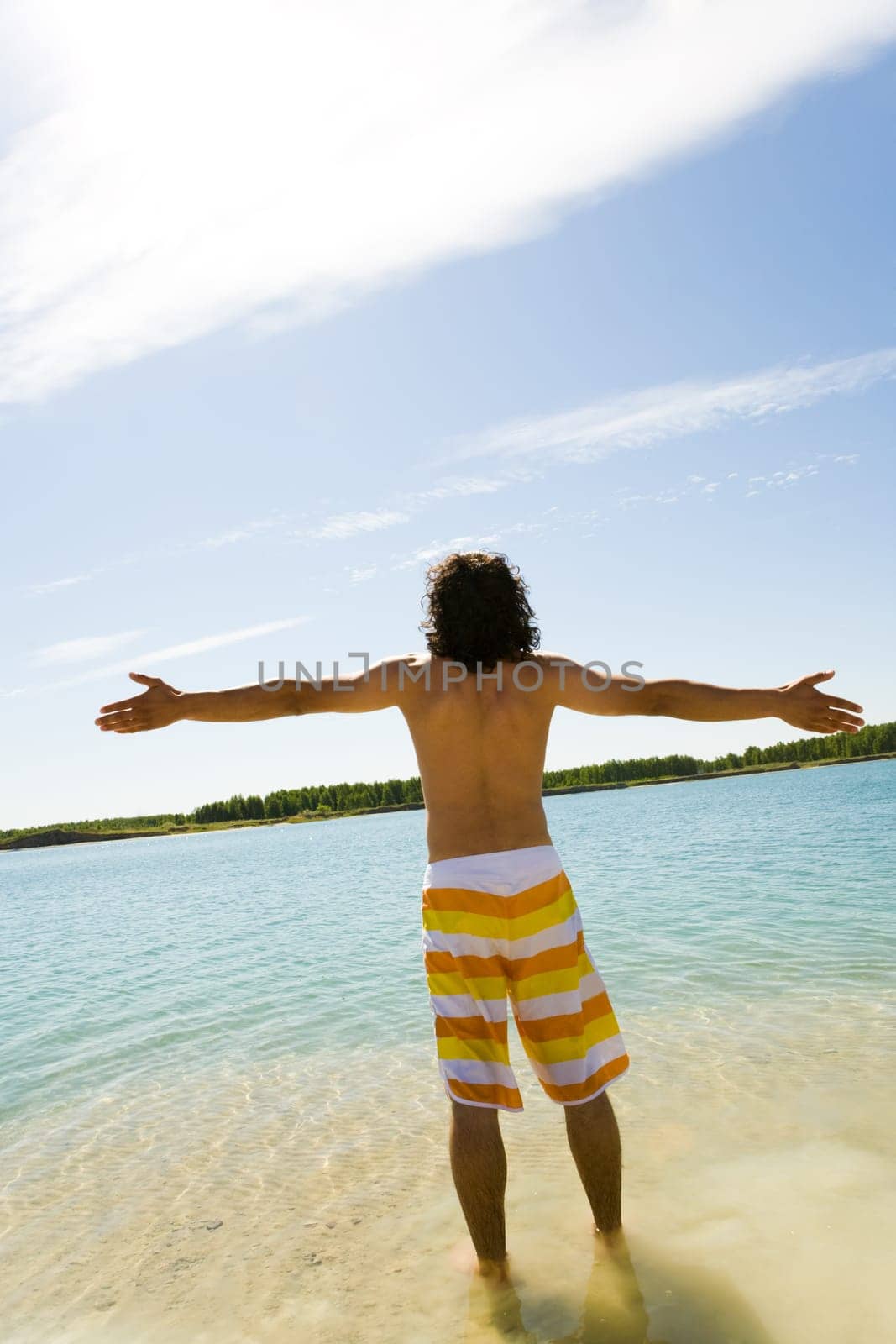 Rear view of young man standing in blue water and having fun with outstretched arms on hot summer day