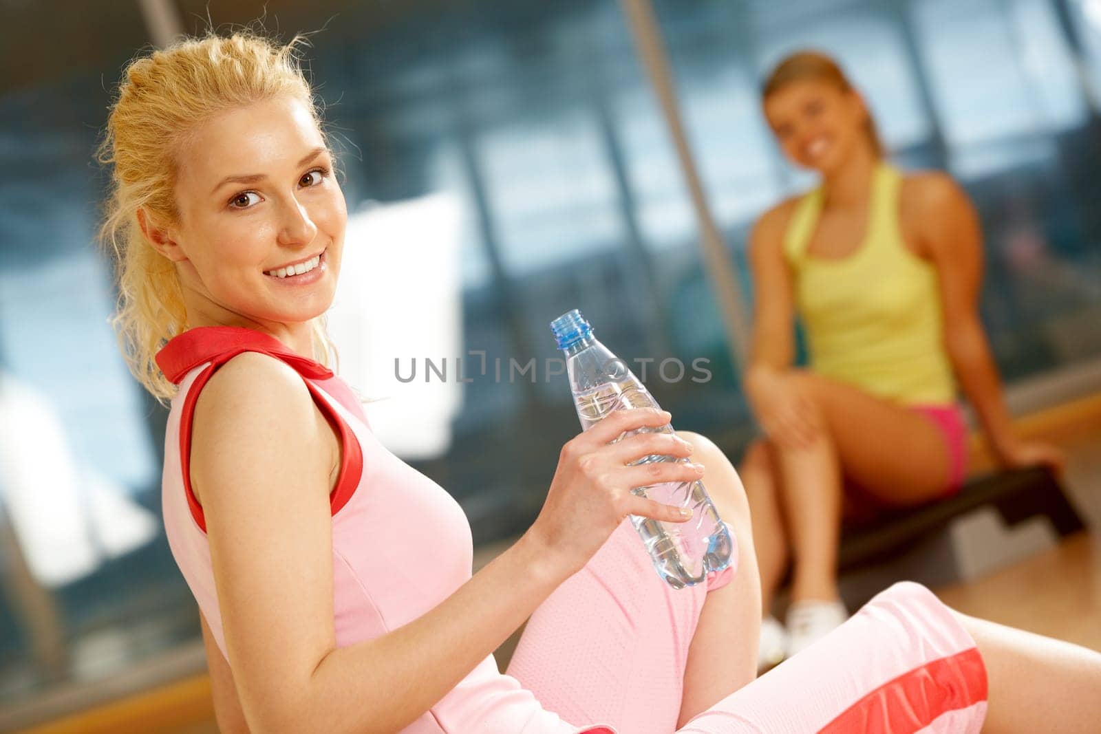 Happy girl sitting on mat with bottle of water with her friend on background