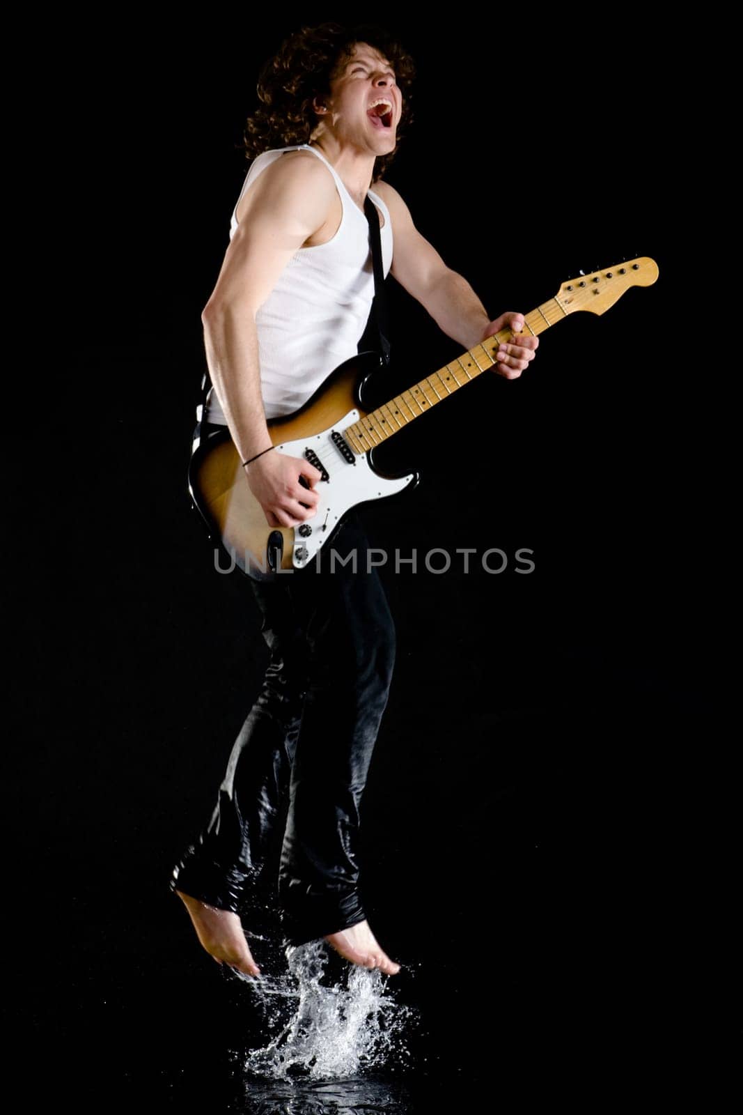 Portrait of dynamic performer playing electrical guitar while jumping above water and singing