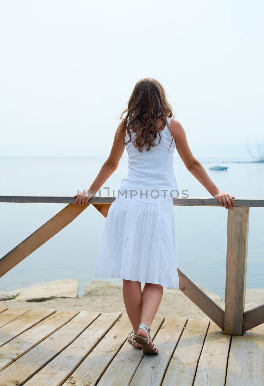 Rear view of pretty young lady in white cotton clothes having rest by the seaside