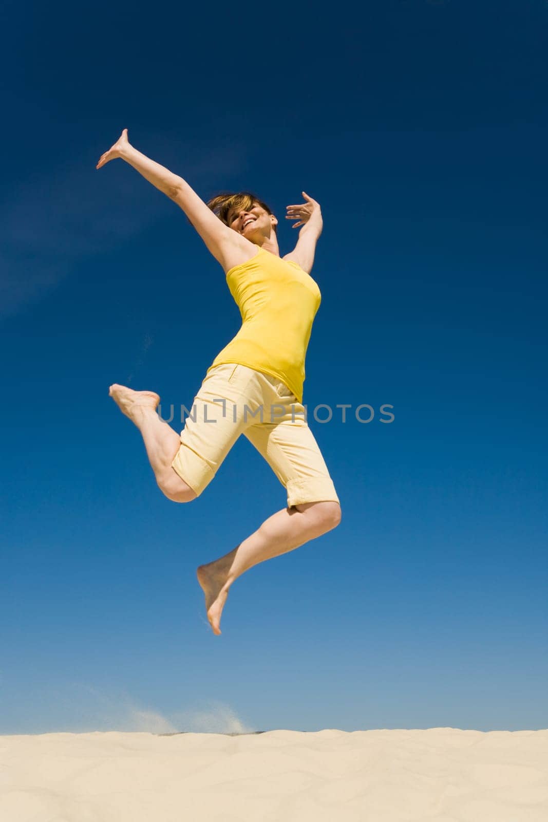 Photo of joyful female in casual clothes jumping over sand with her arms raised