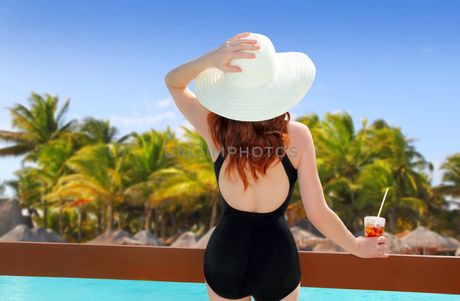 beach hat woman holding cocktail in a tropical beach with rear view black swimsuit