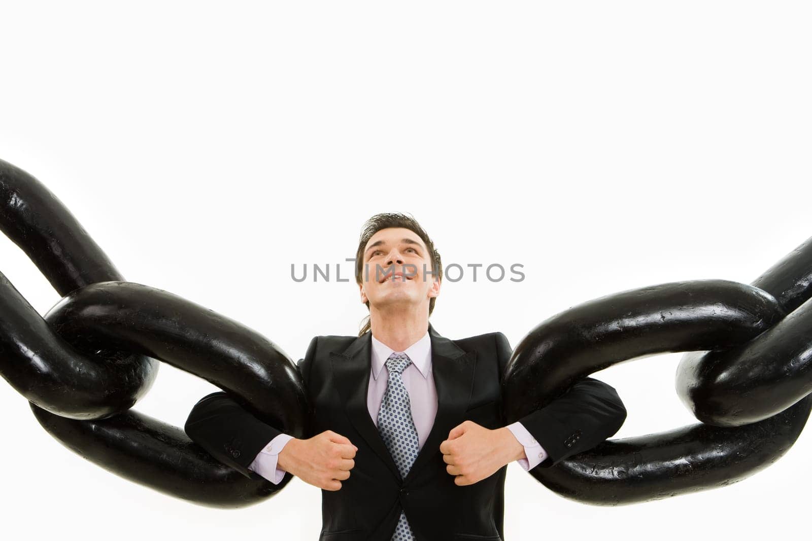 Portrait of powerful businessman holding two sections of huge chain by arms and looking upwards with smile over white background