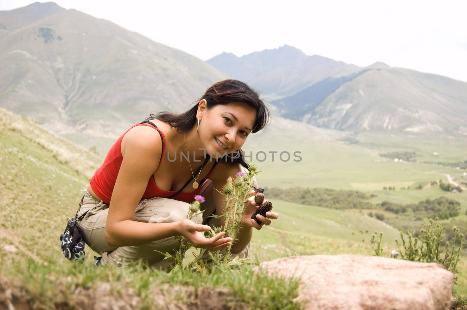 Photo of positive woman with cones in hands touching thorny plants on background of mountains in summer