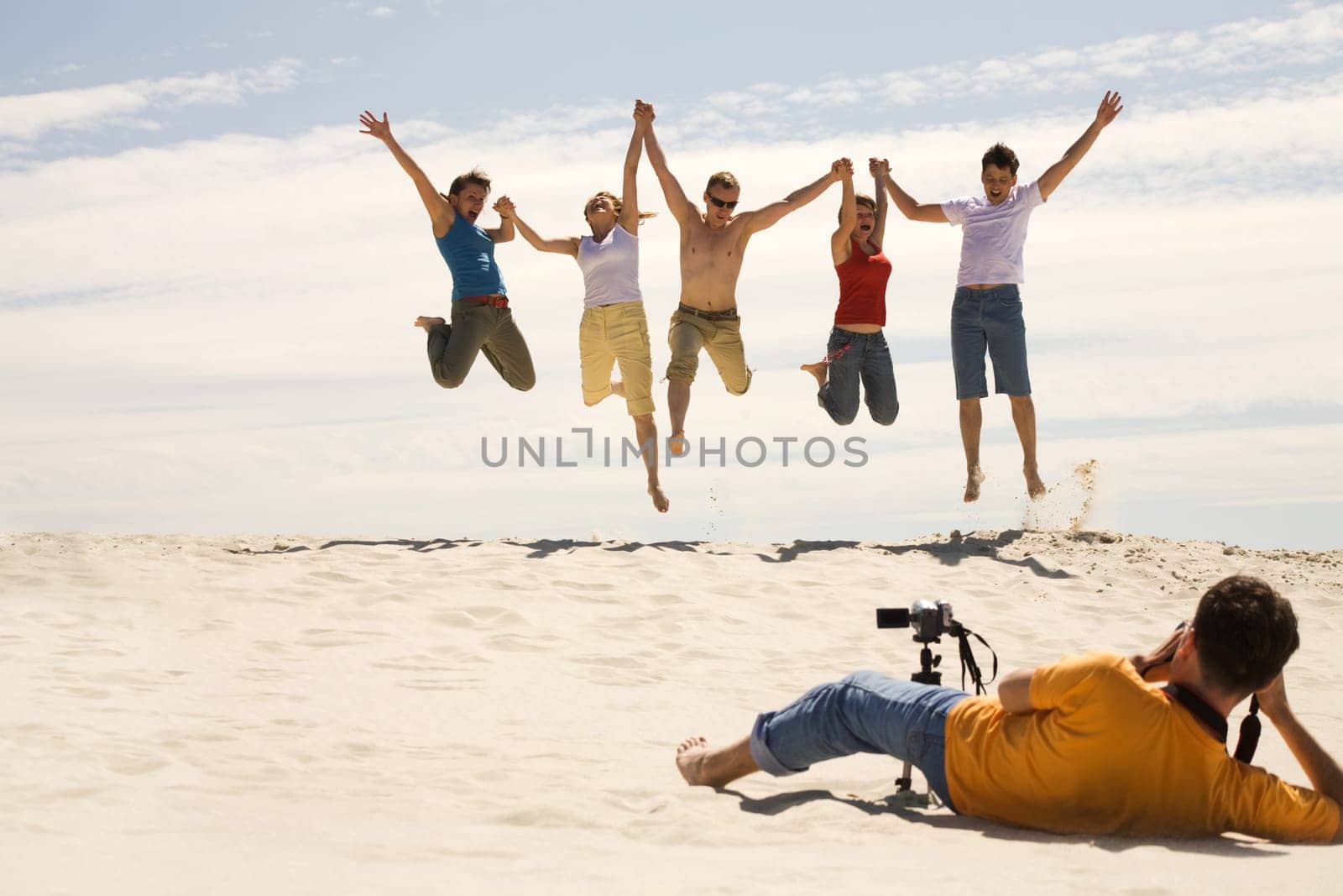 Professional photograpther taking shots of joyful friends jumping on sandy beach against blue sky