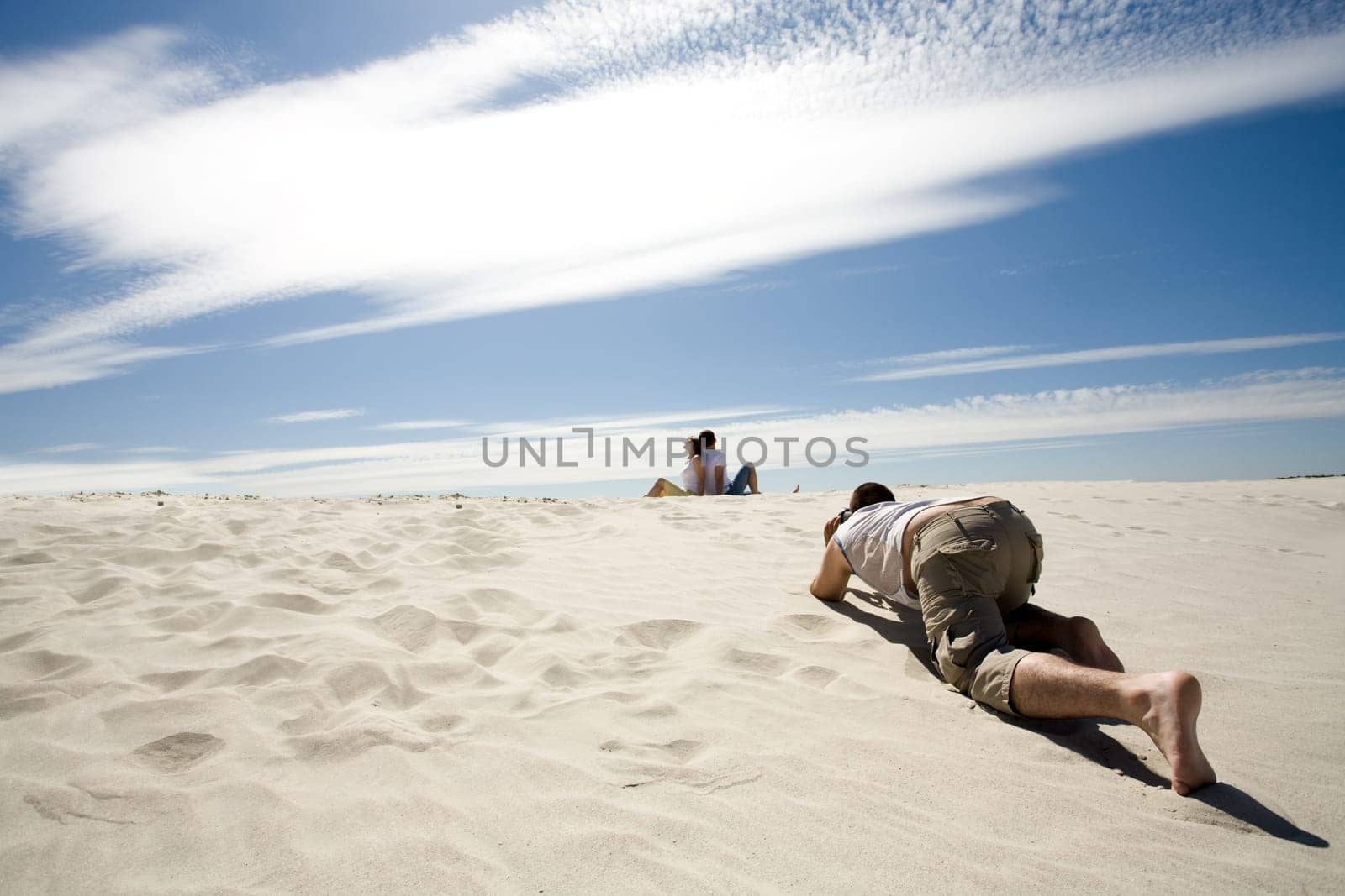 Careful photograpther taking shots of relaxing couple sitting back to back on sandy beach against blue sky