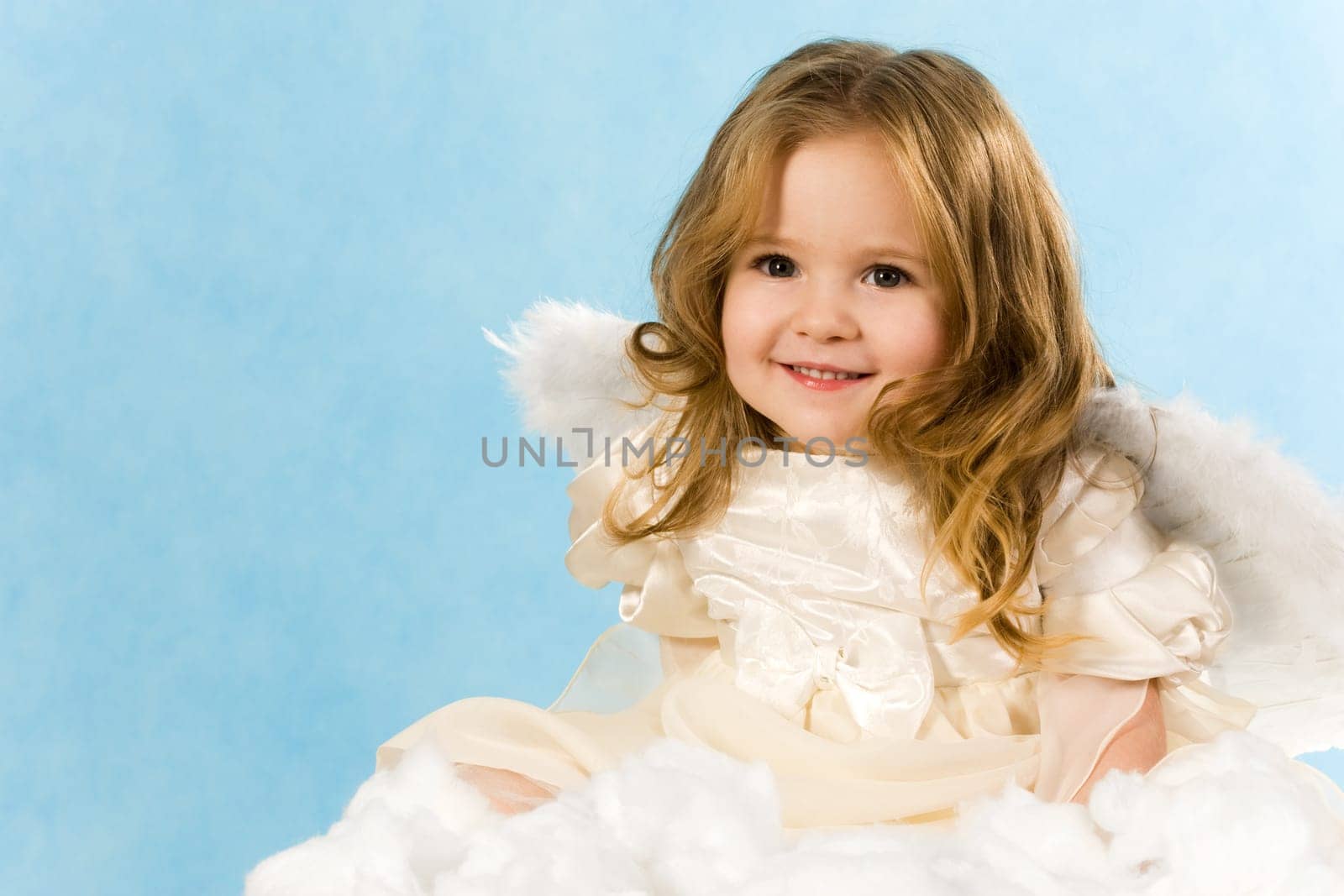 Happy little girl wearing angel dress sitting on white cloud and smiling