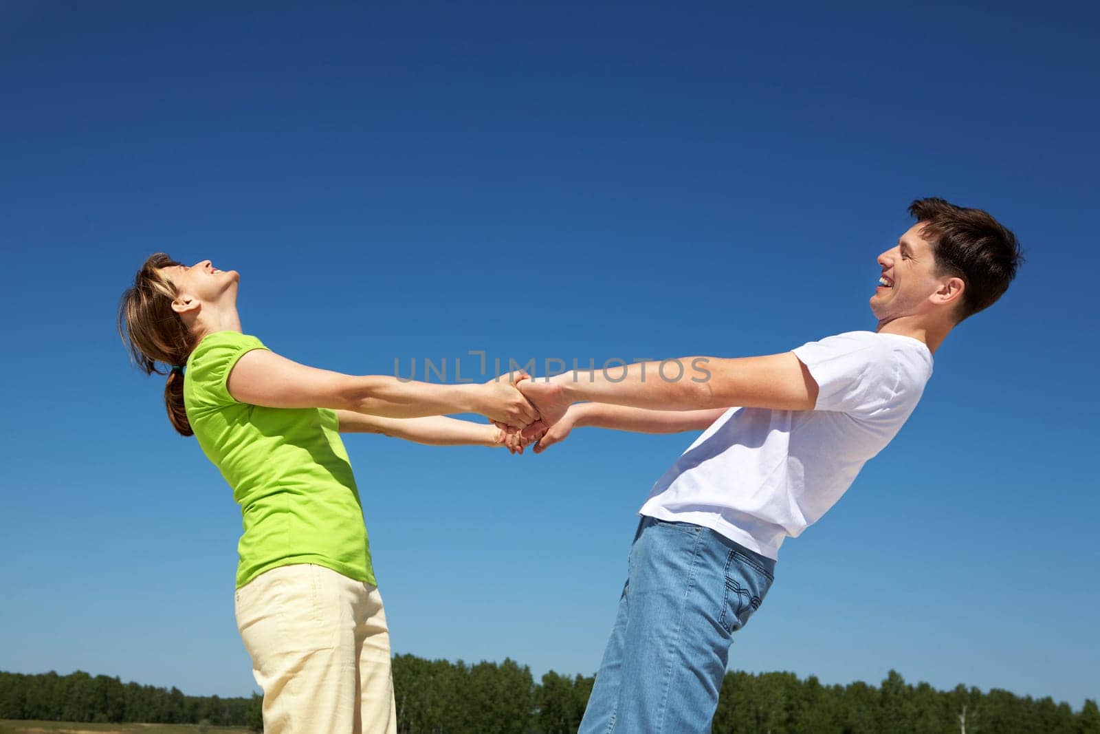 Photo of playful couple holding each other by hands with their arms stretched against blue sky
