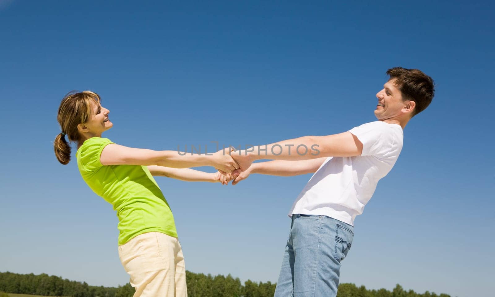 Photo of playful couple holding each other by hands with their arms stretched against blue sky