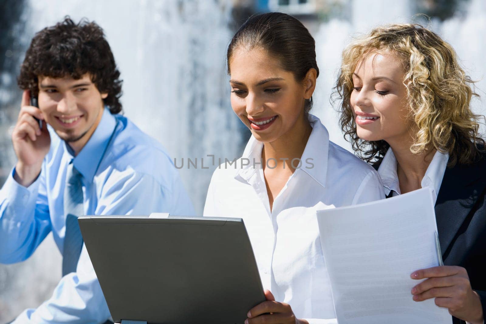 Two cute smiling female students are reading documents outside and handsome young man talking by the phone staring at them