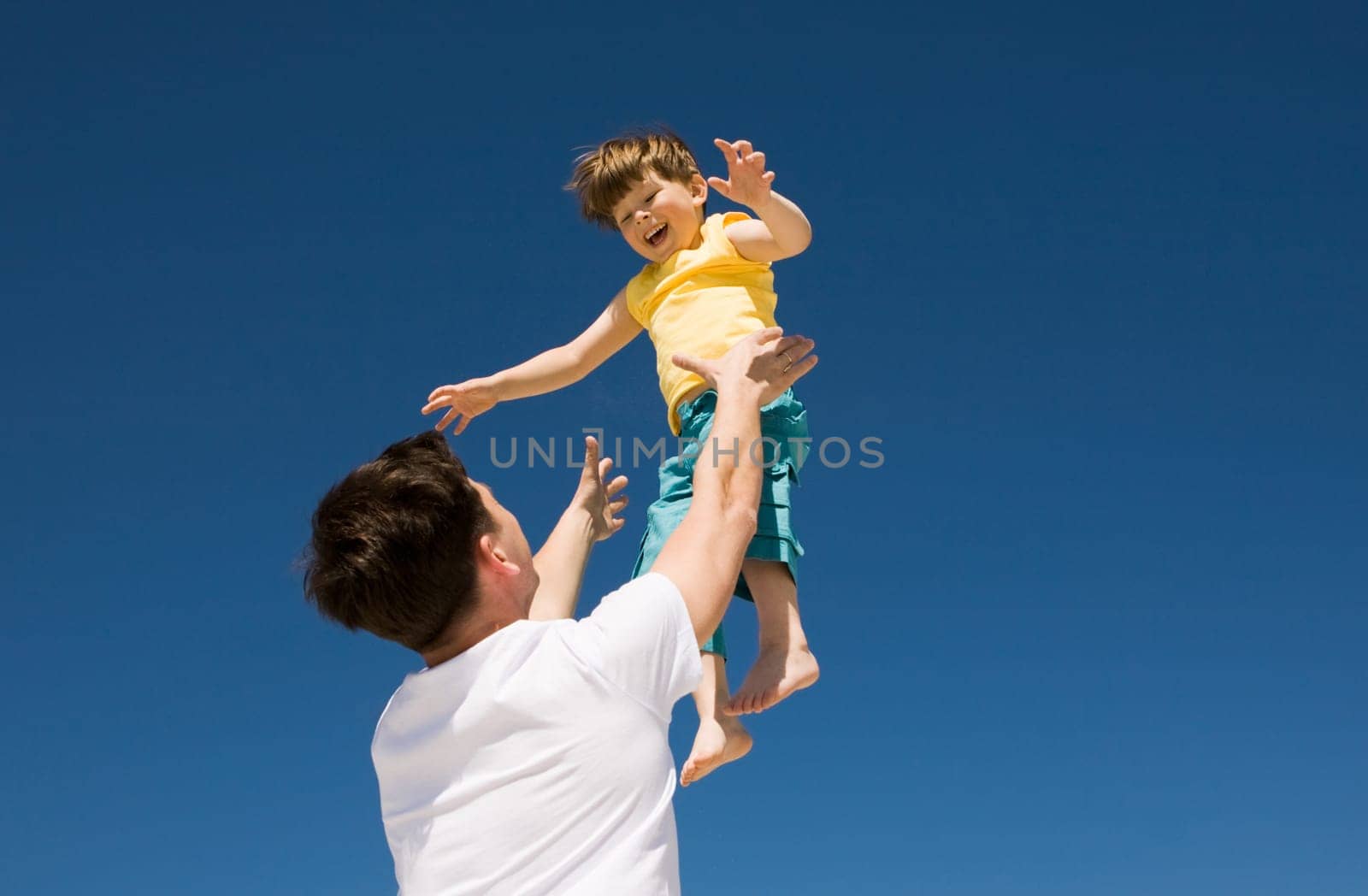 Photo of happy man throwing his cute son up with bright blue sky at background