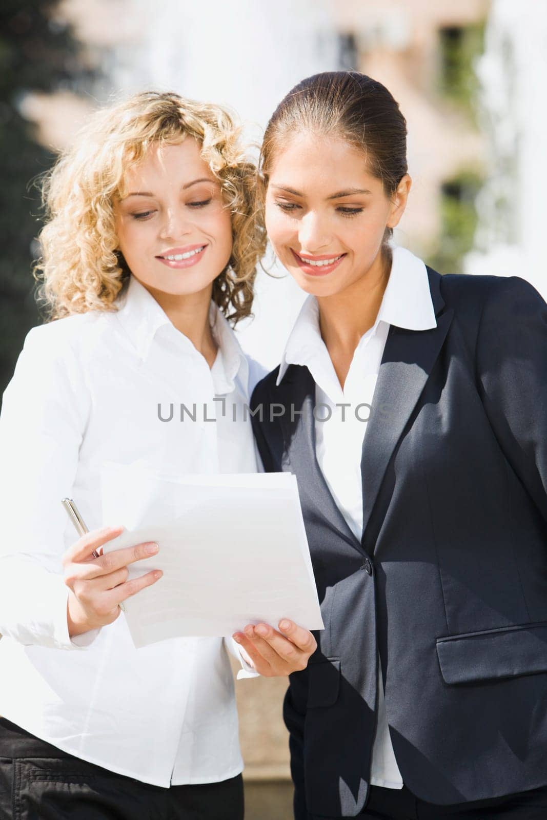 Two cute smiling female students standing outside ahead of the fountain reading the documents