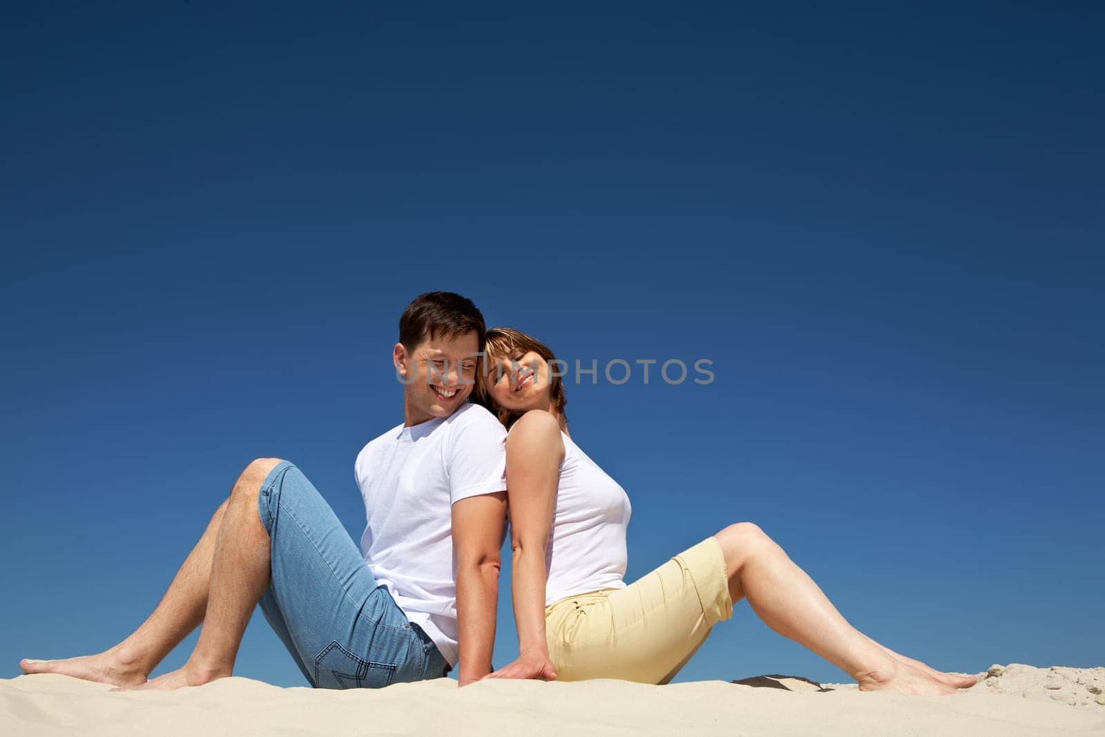 Portrait of relaxing couple sitting back to back on sandy beach against blue sky