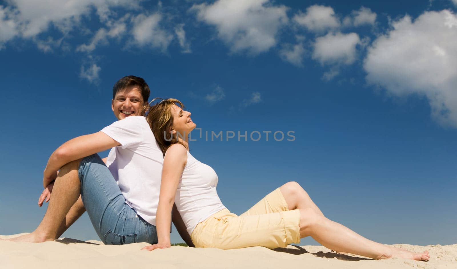 Portrait of relaxing couple sitting back to back on sandy beach against blue sky