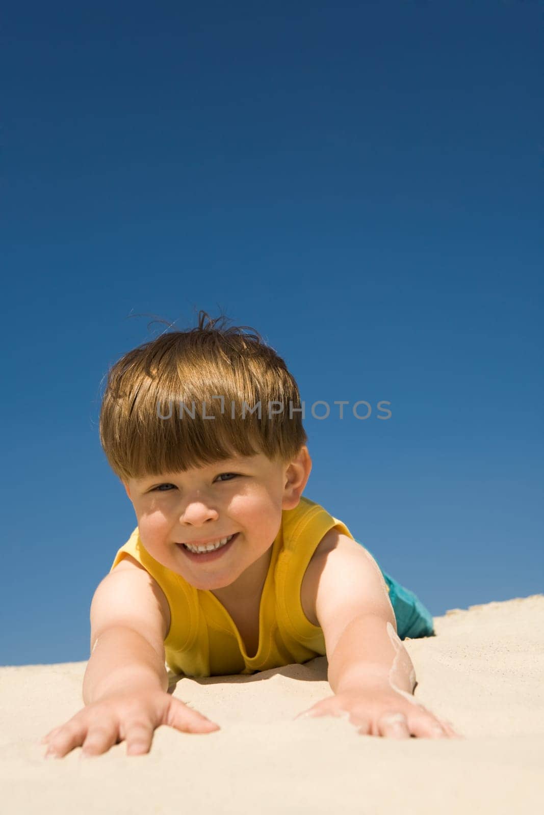 Happy boy lying on sand and looking at camera with smile during summer vacations