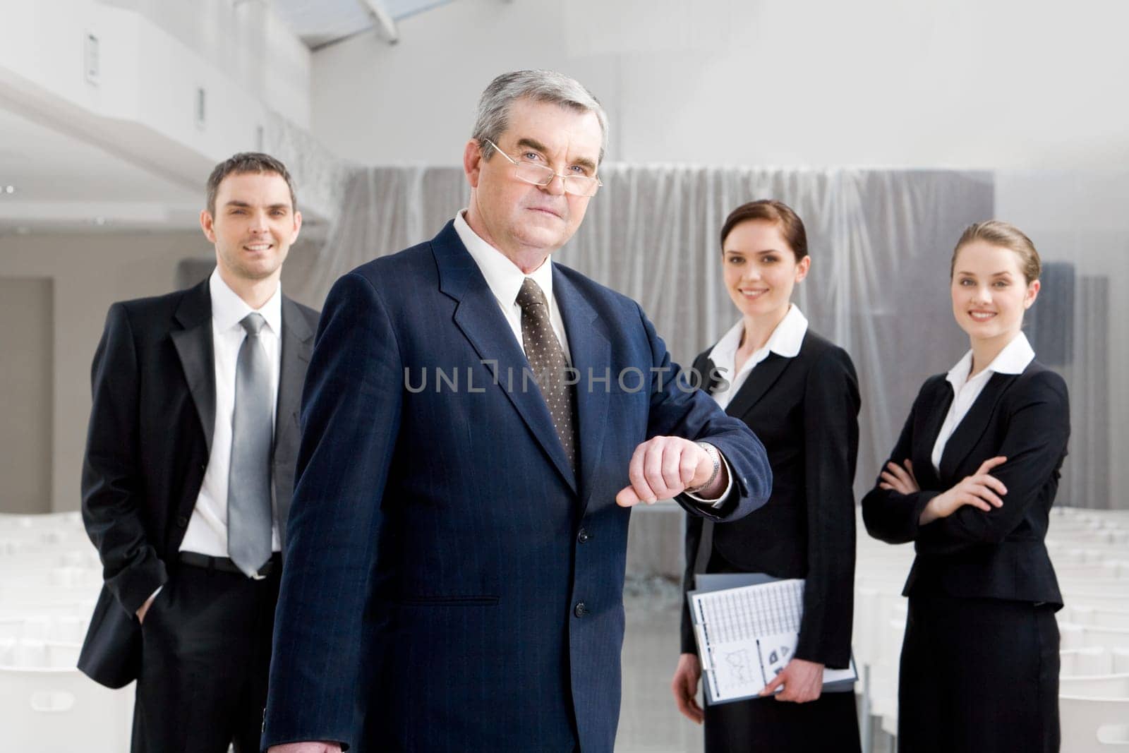 Portrait of senior businessman looking at camera while watching the time at background of confident managers