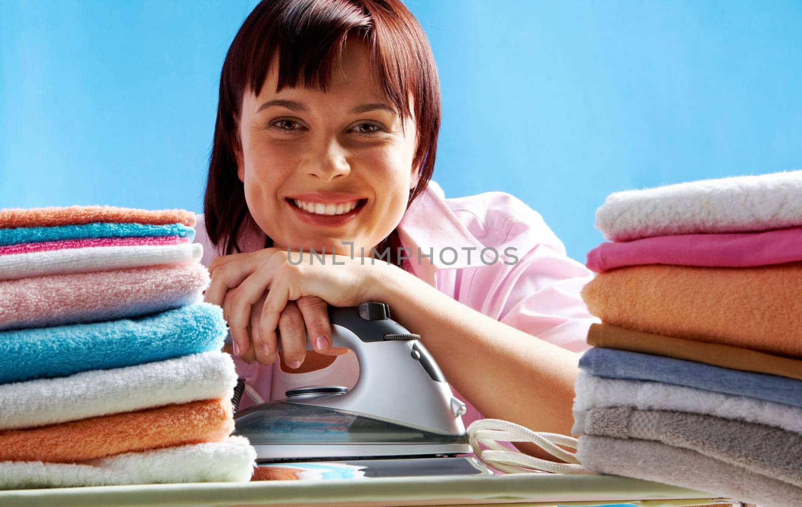 Portrait of young female with iron between two stacks of colorful towels smiling at camera