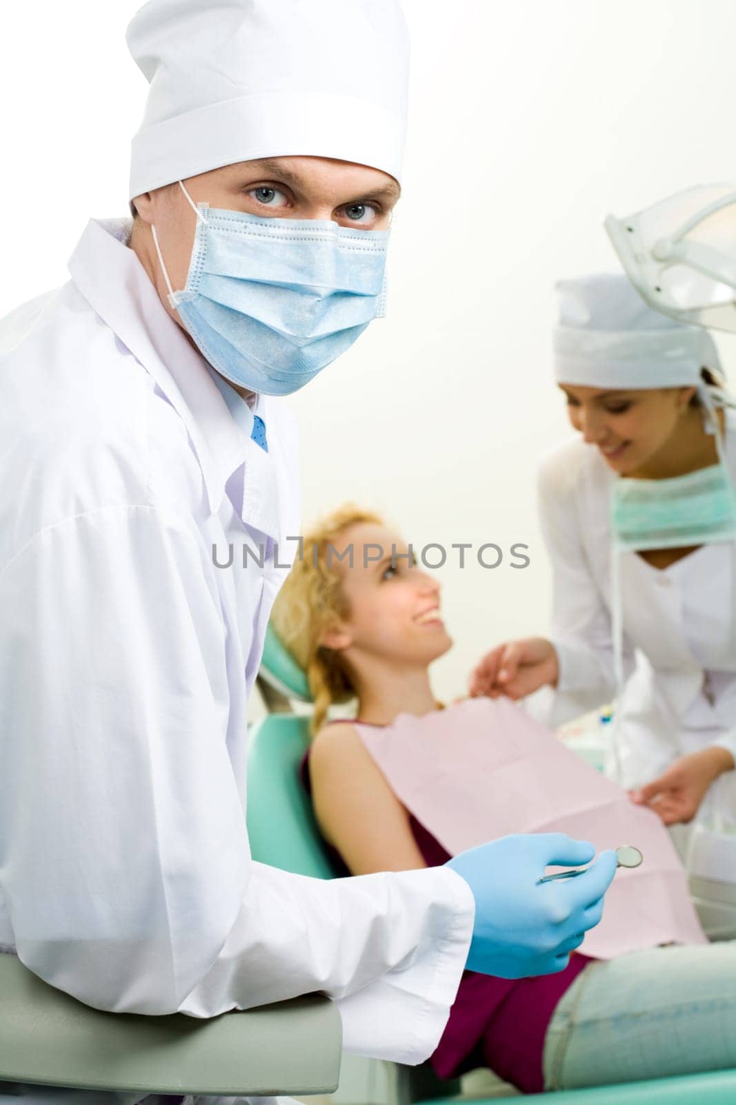 Portrait of confident doctor in sterile mask on background of his assistant helping patient