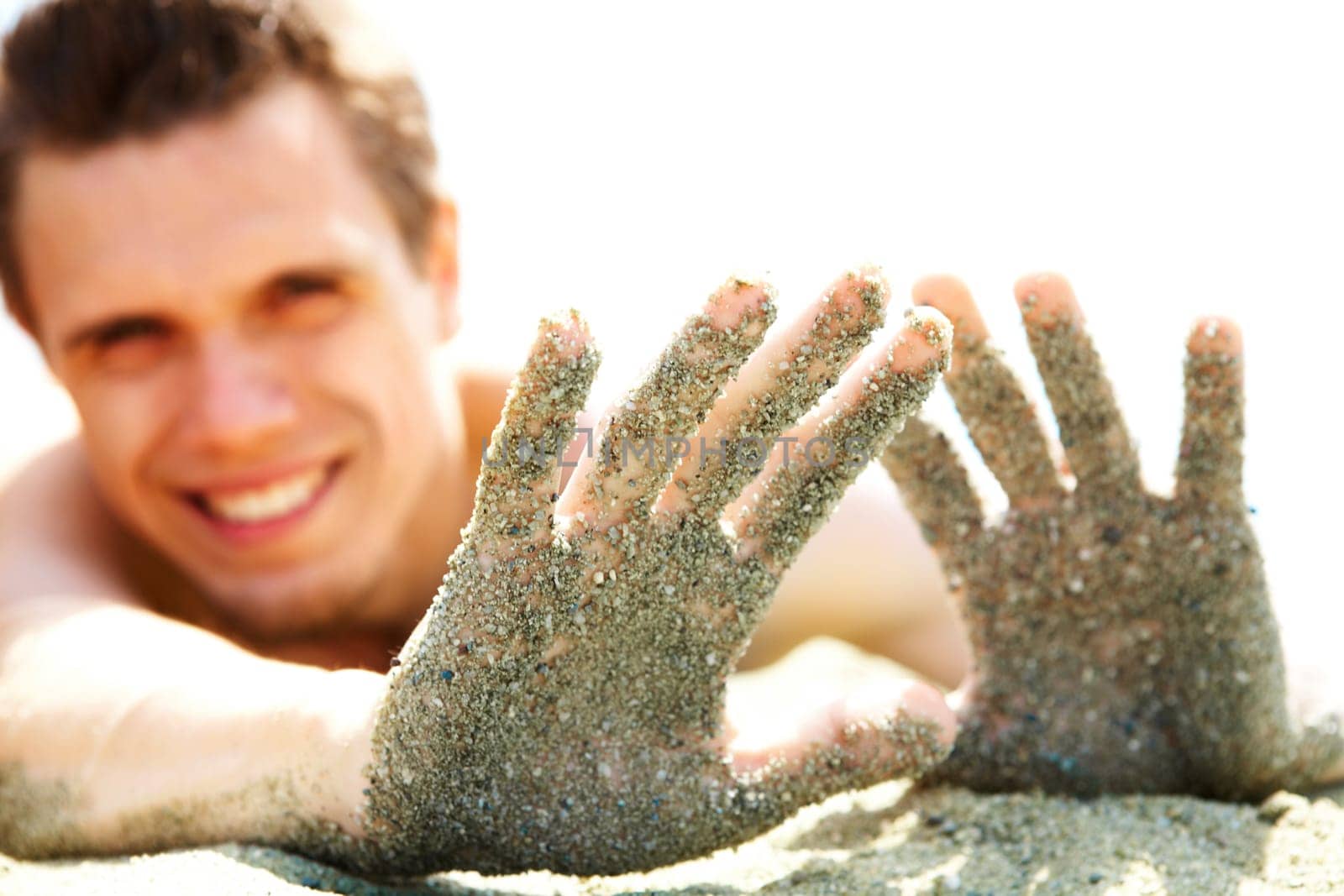 Photo of smiling guy lying on beach and showing his hands covered with sand while sunbathing