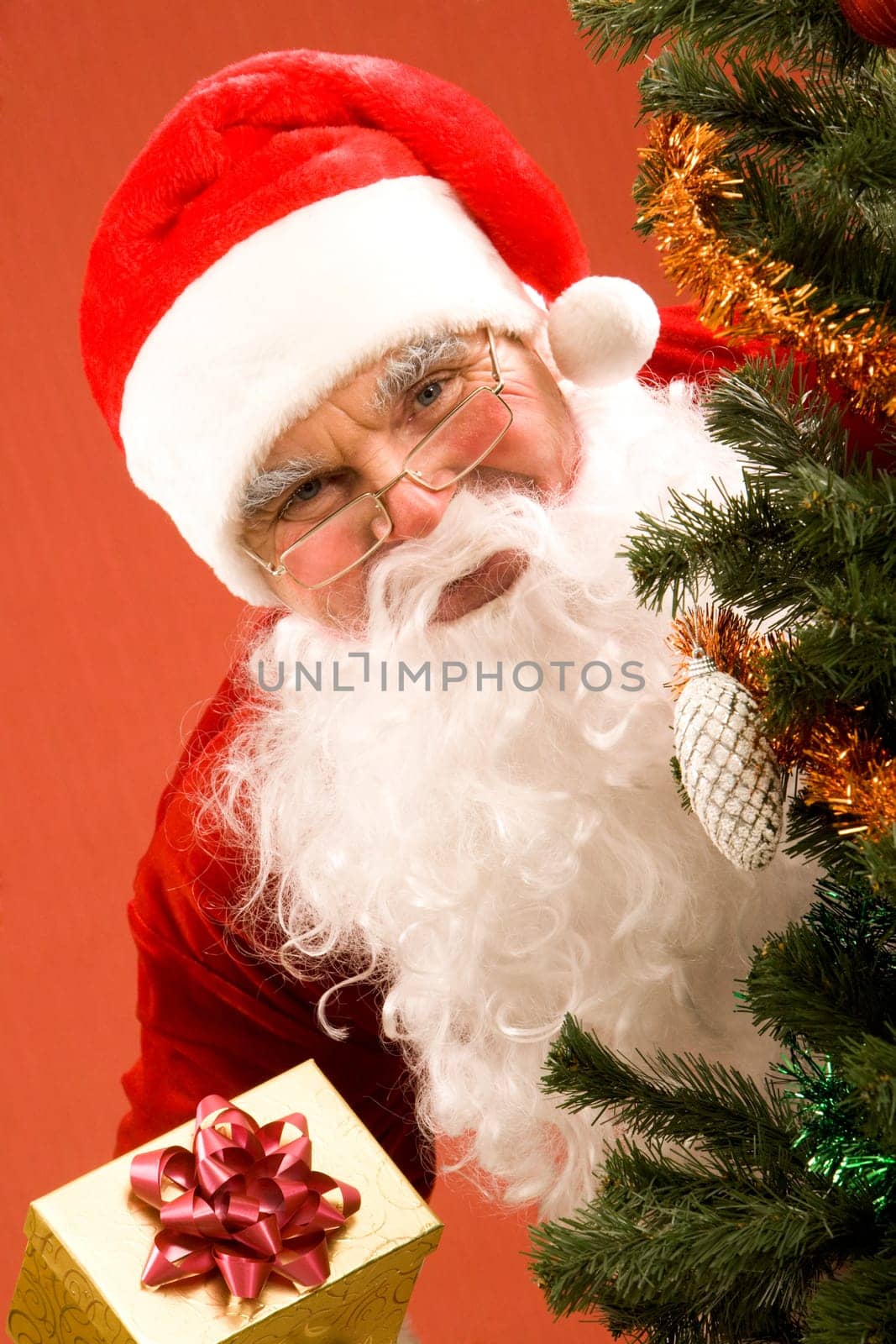 Photo of happy Santa Claus with small giftbox looking at camera out of decorated xmas tree