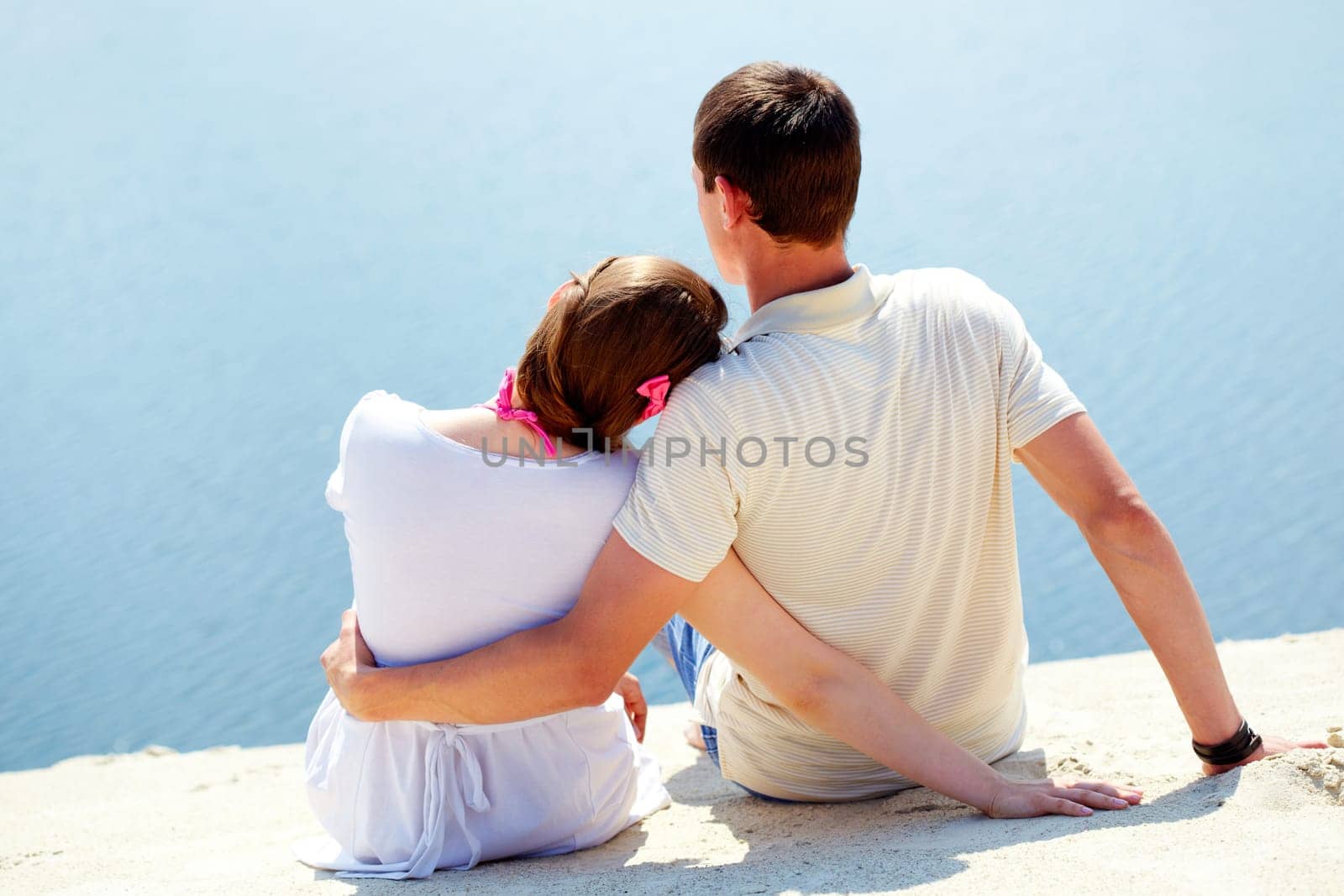 Backs of peaceful couple sitting on sandy shore in front of blue water