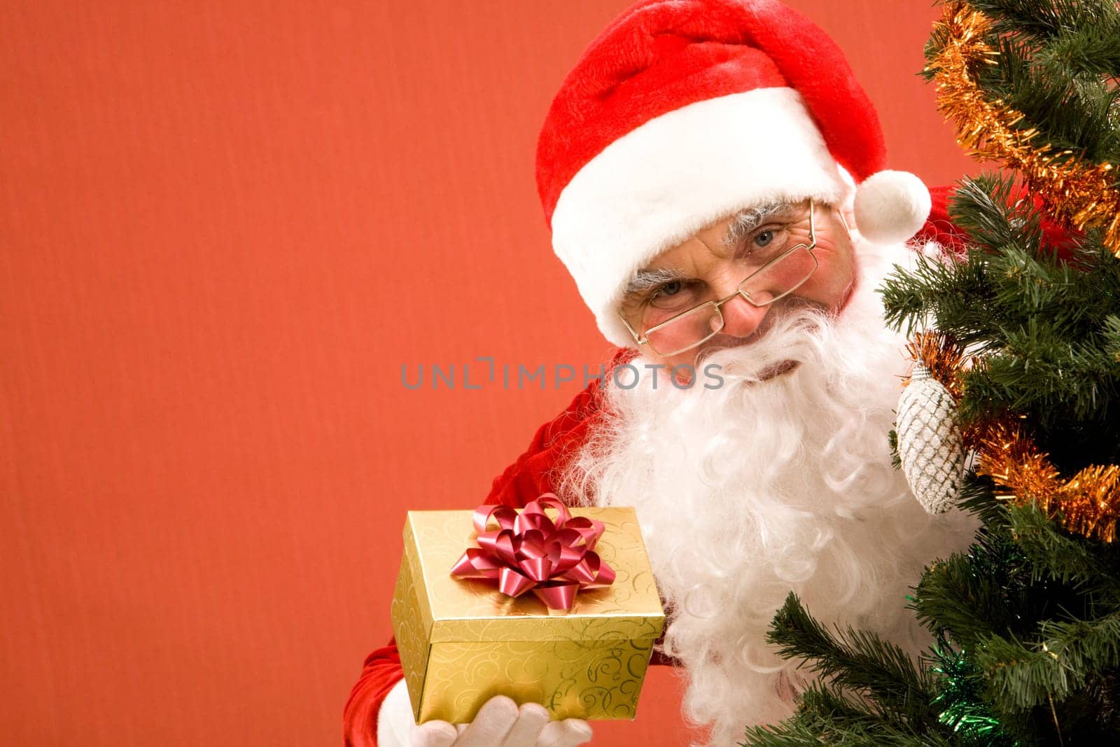 Photo of happy Santa Claus with small giftbox looking at camera out of decorated xmas tree