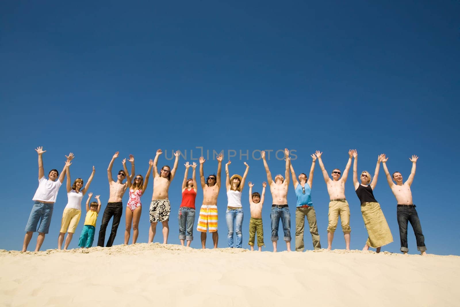 Image of many friends standing on sandy beach with their arms raised against blue sky