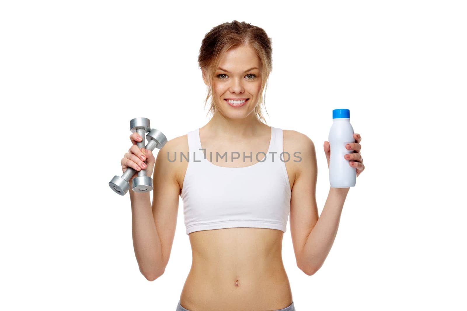 Portrait of a slim girl holding a bottle of milk and dumbbells, looking at camera and smiling