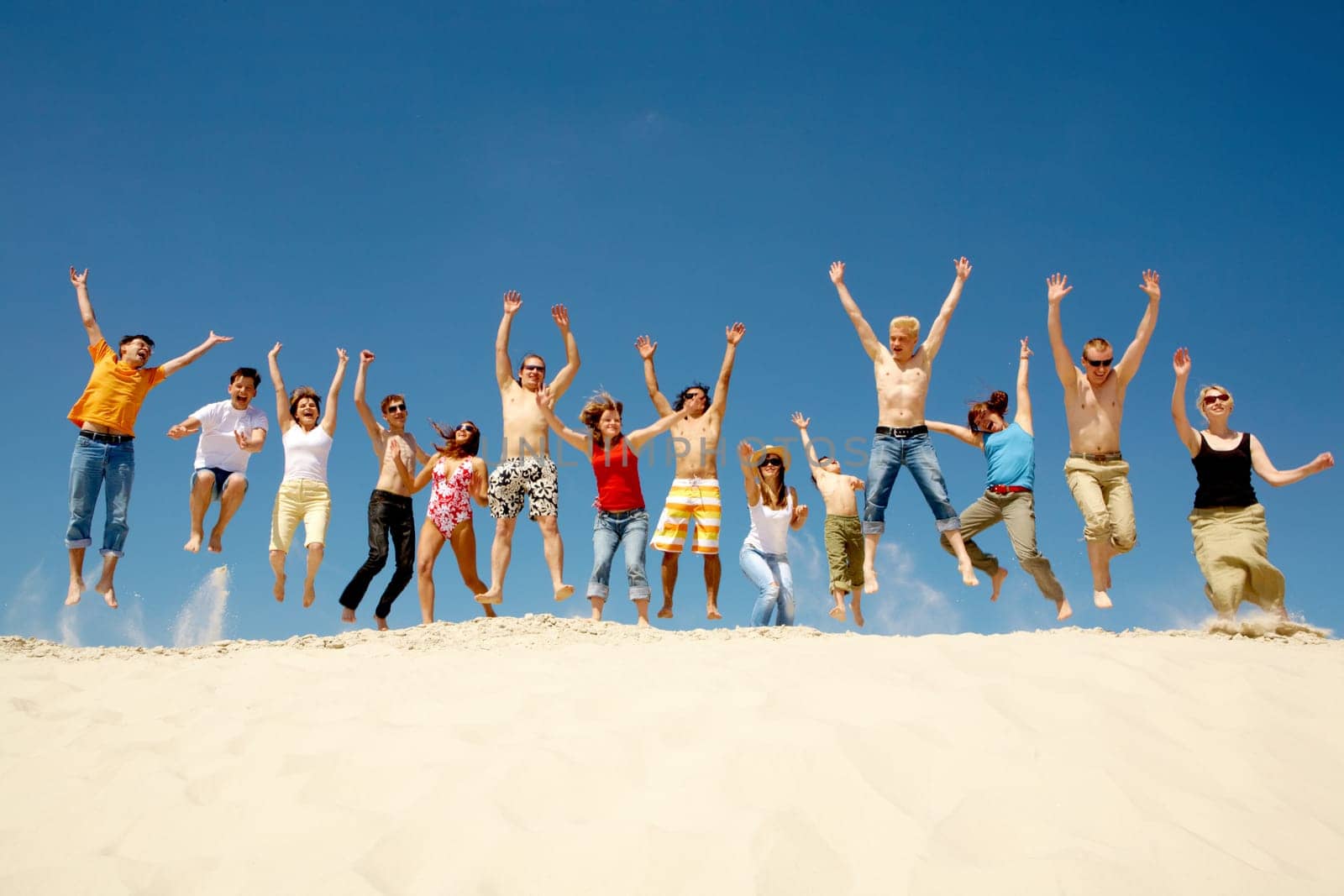 Crowd of friends jumping on sandy beach with their arms raised against blue sky