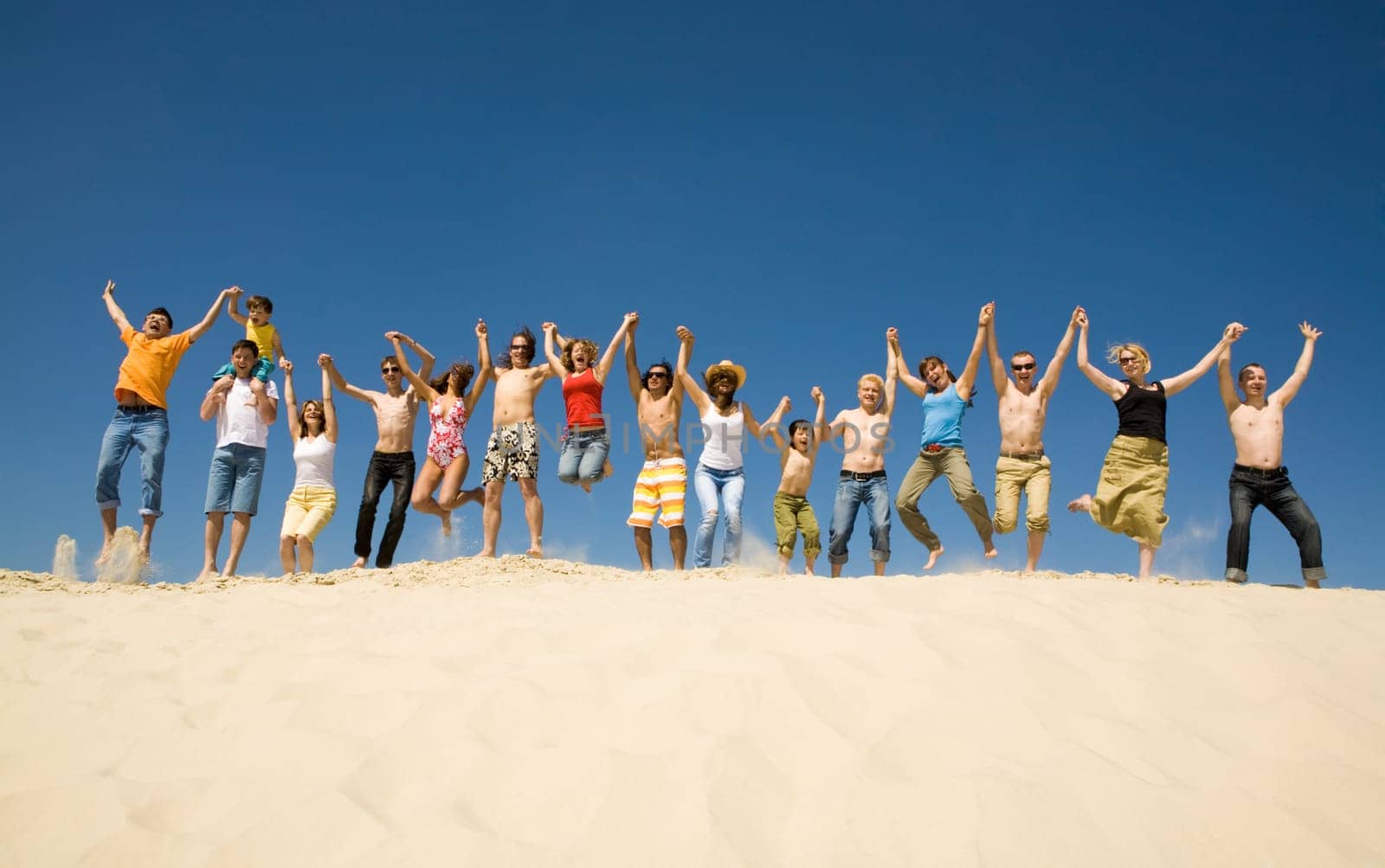 Crowd of friends jumping on sandy beach with their arms raised against blue sky