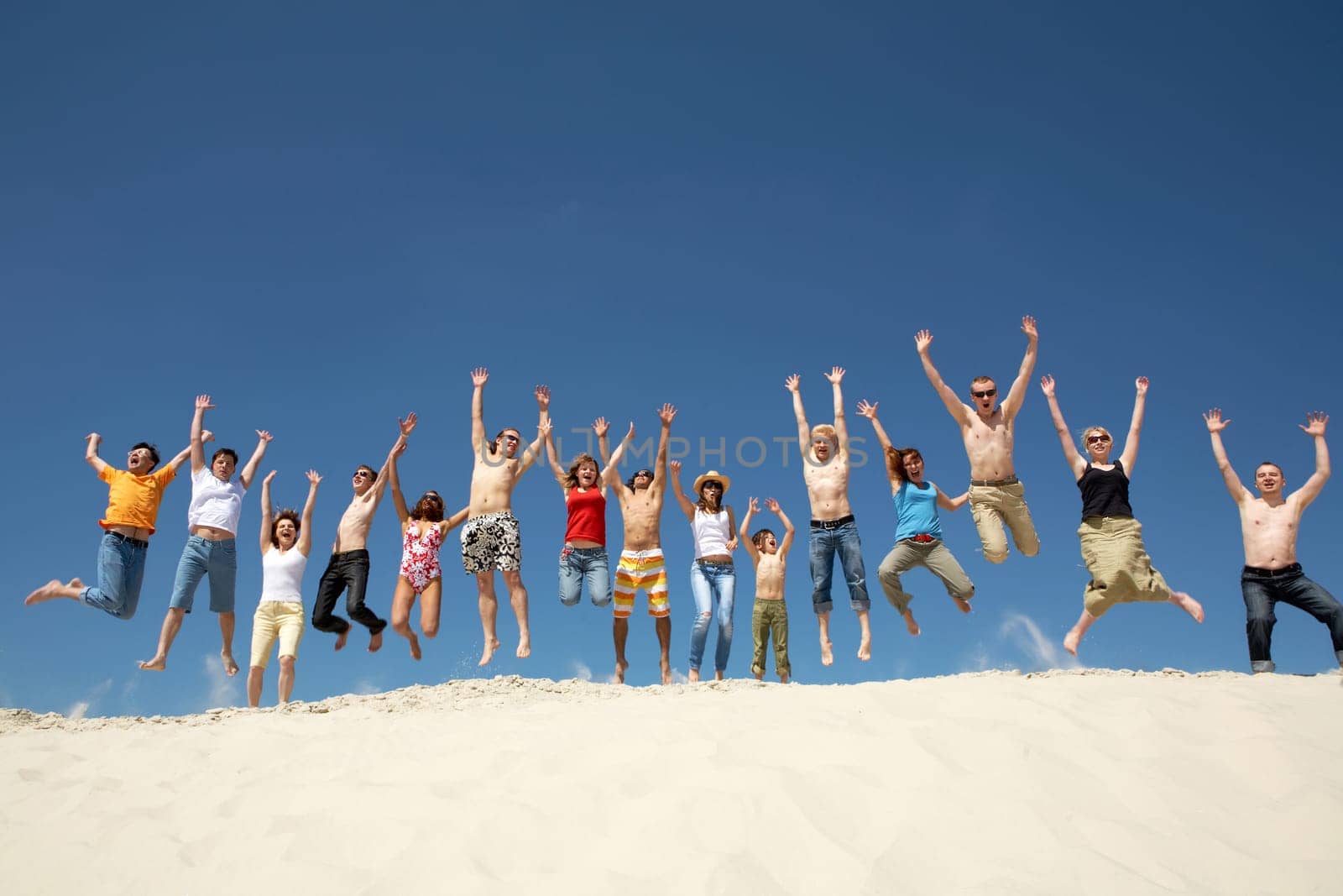 Crowd of friends jumping on sandy beach with their arms raised against blue sky