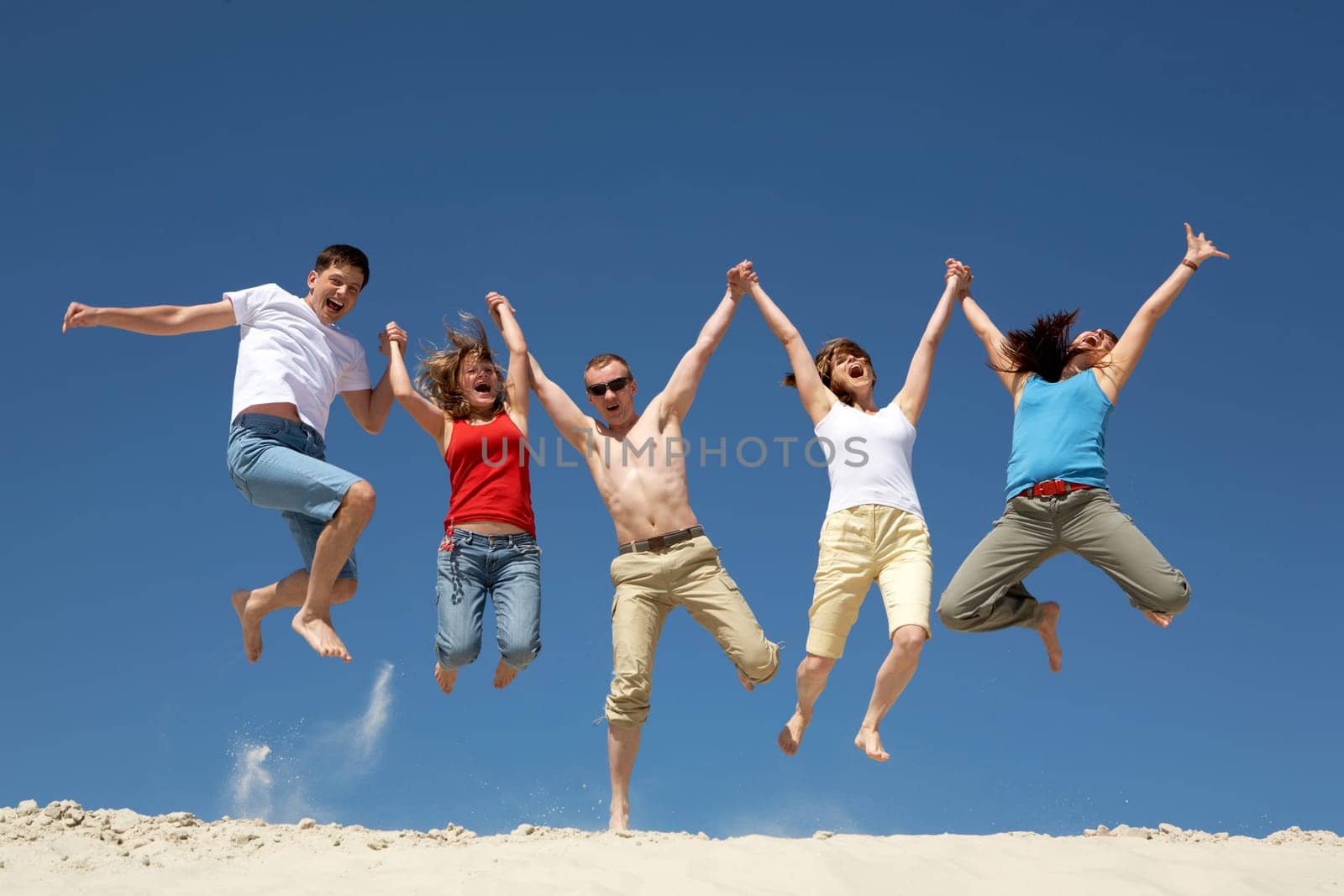 Photo of excited people jumping on sandy beach with their arms raised against blue sky