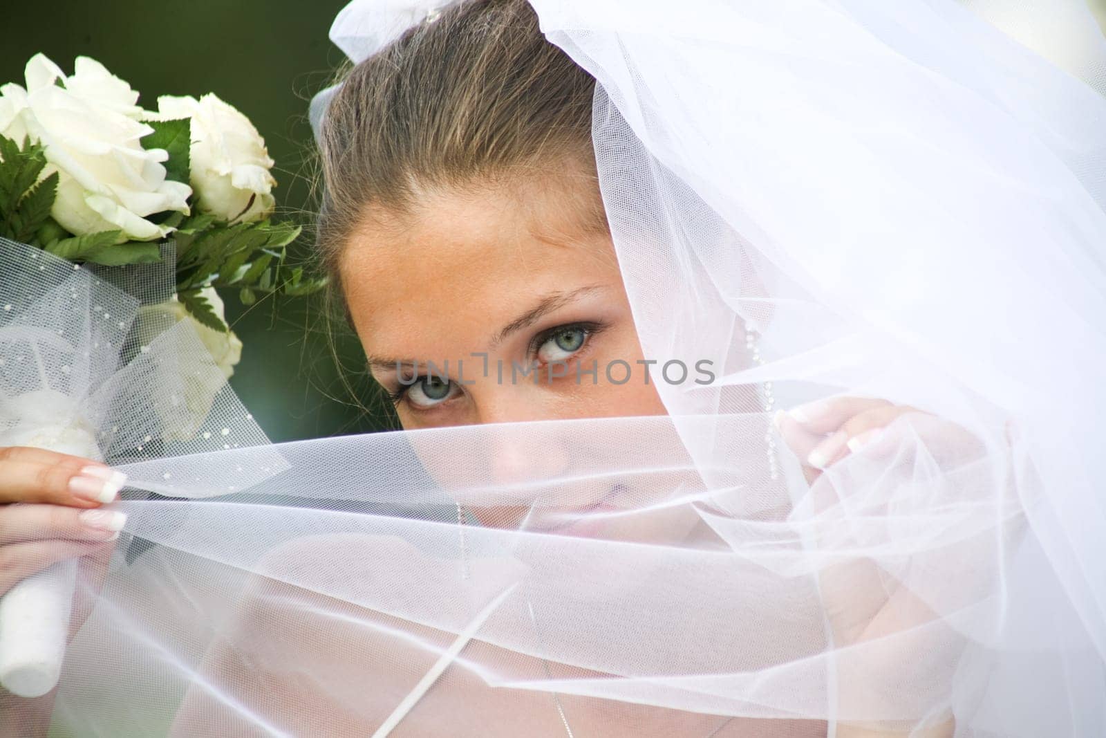 Close-up of young green-eyed bride holding veil by her face and looking at camera